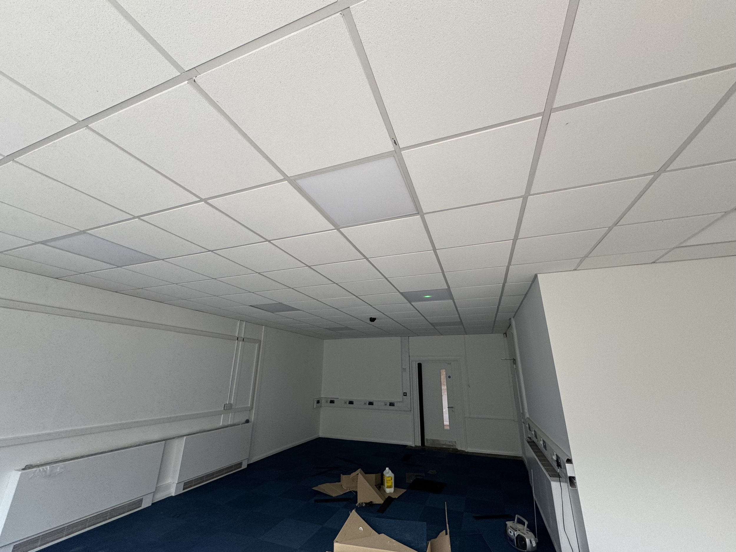 Ceiling with white acoustic tiles, walls with baseboards, and scattered boxes and cleaning supplies on the dark blue carpeted floor.