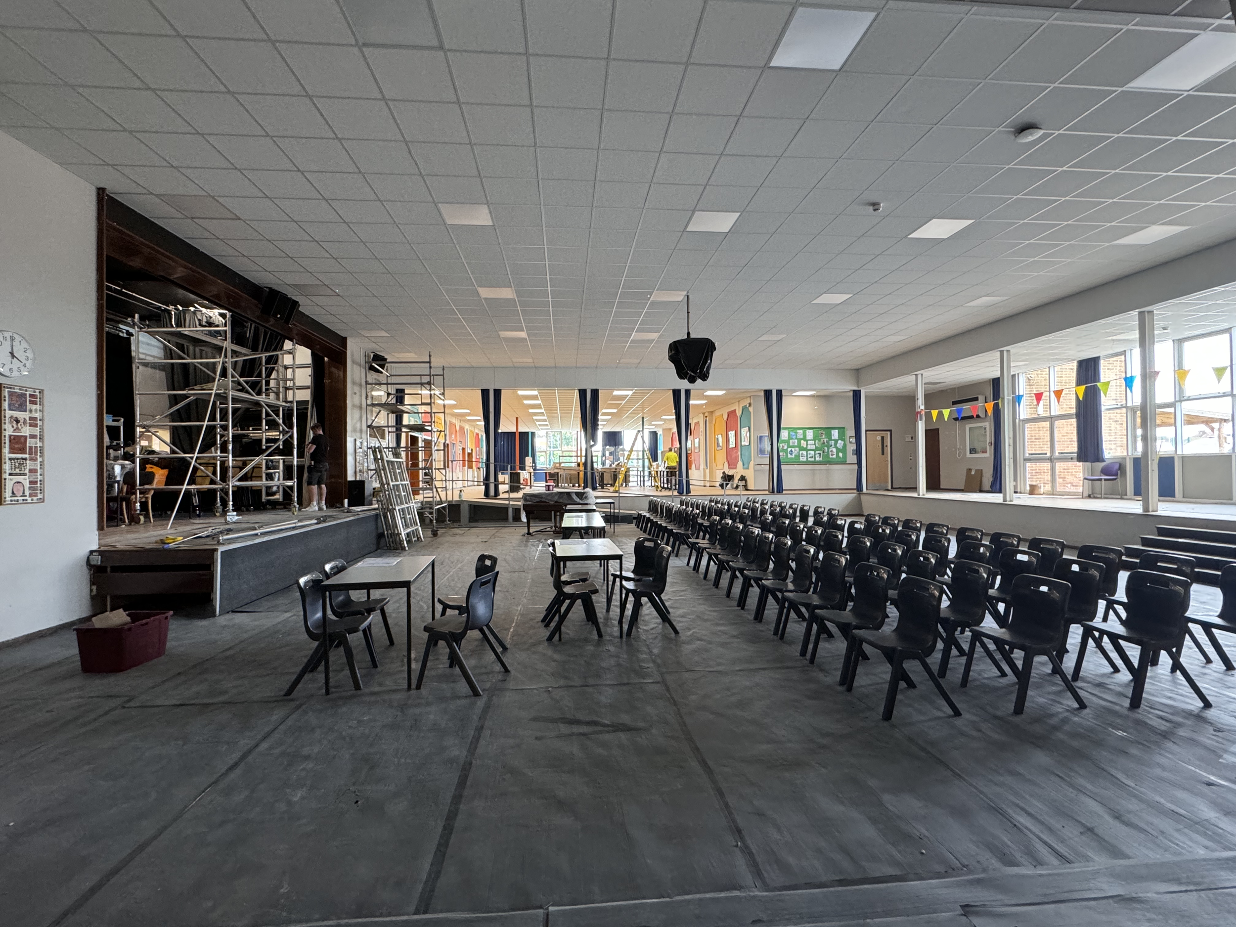 An indoor space with black chairs arranged in rows. Construction or renovation work is underway with scaffolding, ladders, and workers, suggesting the space is being prepared or repaired.