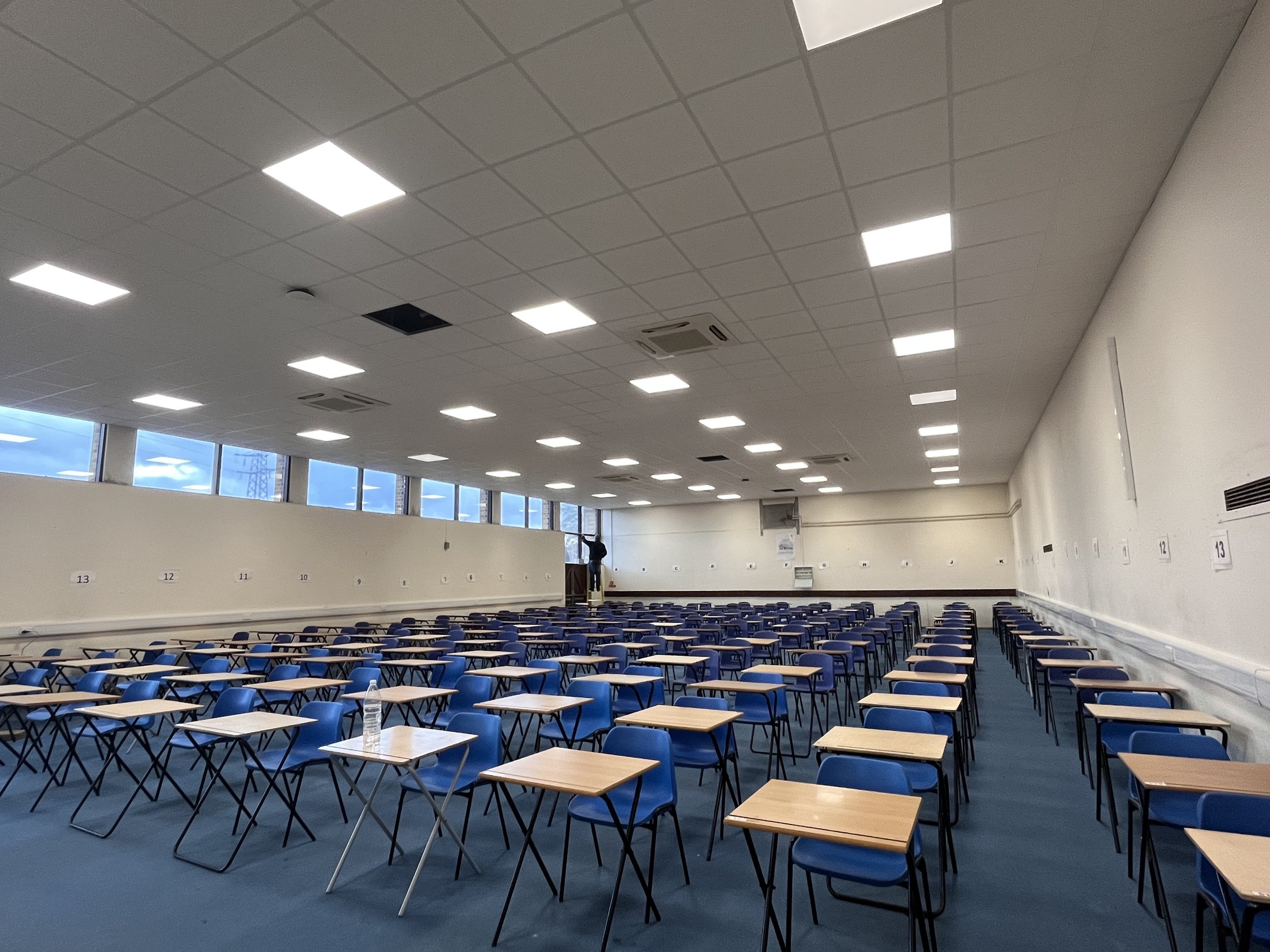 a school hall with rows of blue chairs and wooden desks, white walls, and ceiling lights, with large windows showing a cloudy sky outside.