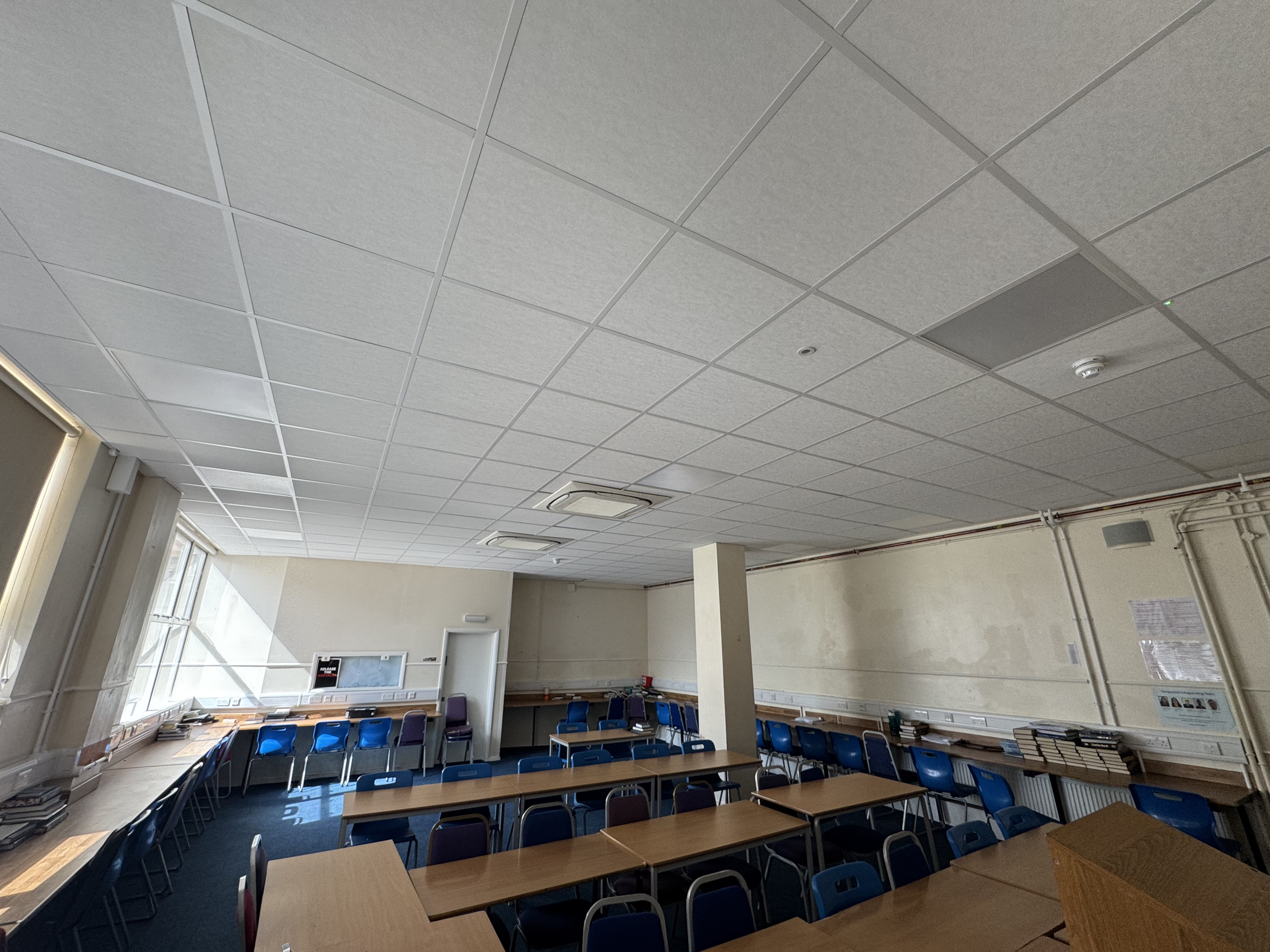 An empty classroom with wooden tables and blue chairs, high ceilings with suspended ceiling tiles, windows along the wall, and books stacked on tables and shelves.