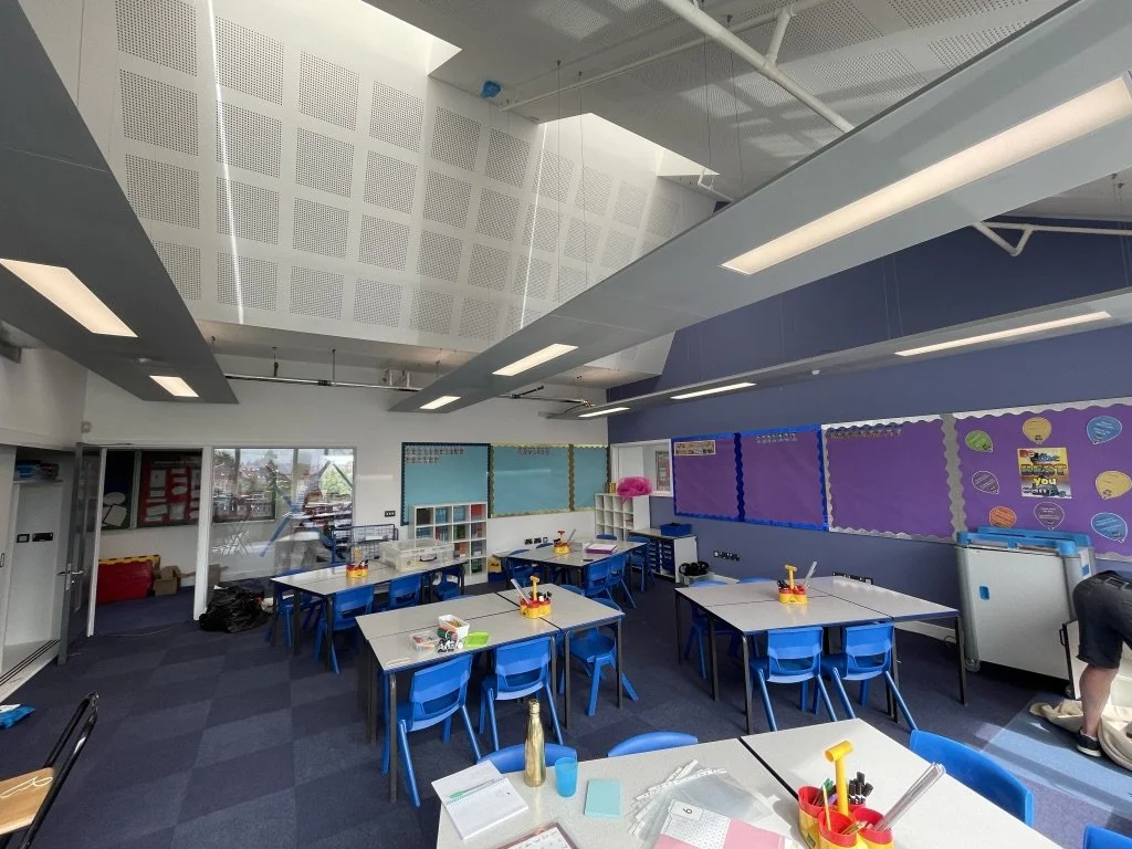 Empty classroom with white tables and blue chairs, decorated bulletin boards on the walls, and large windows letting in natural light.