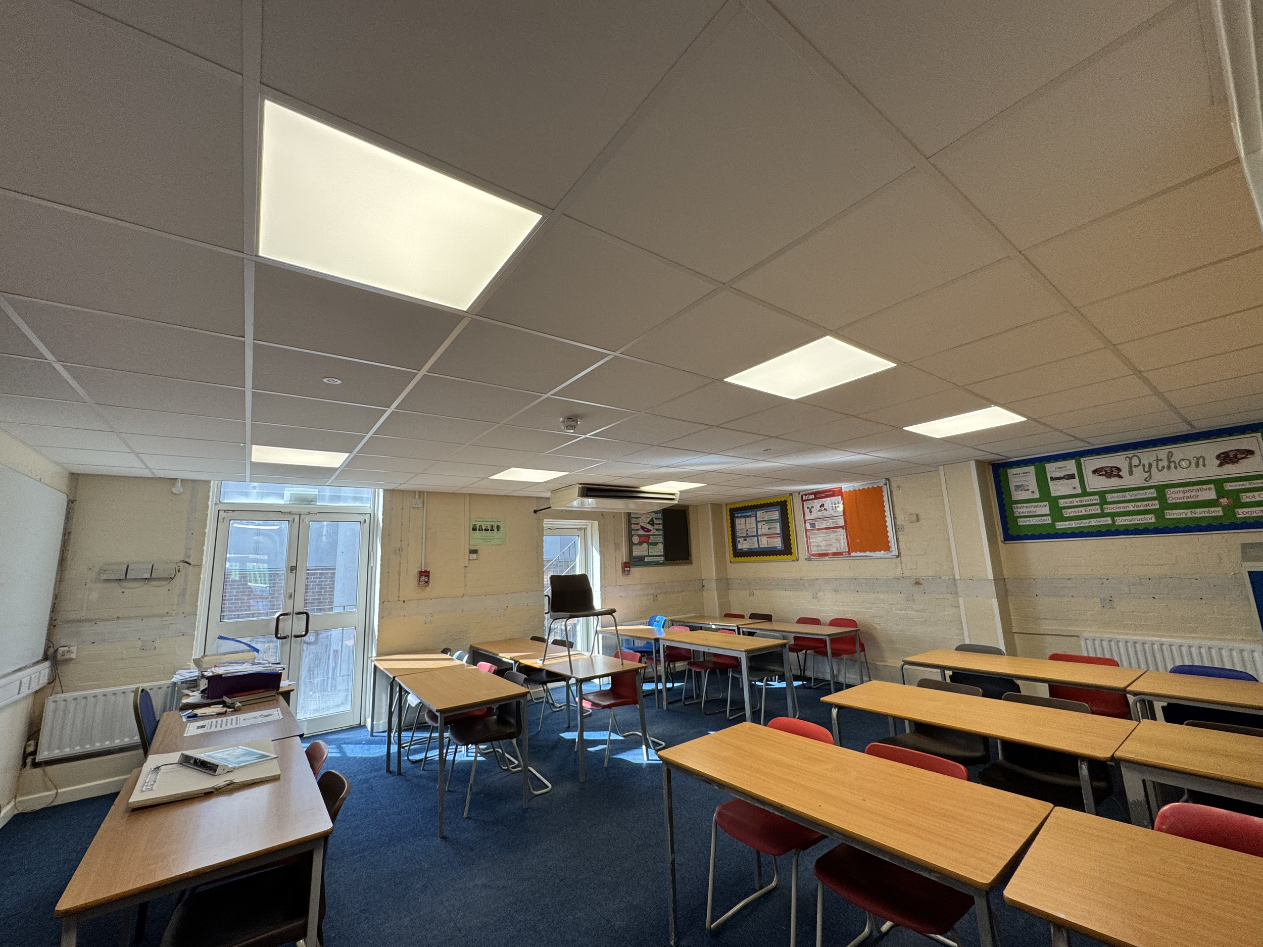 Classroom with rows of desks and chairs, windows with blinds, educational posters on the walls, and ceiling lights.