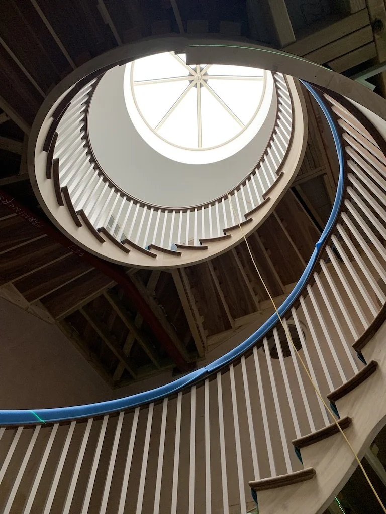 A winding wooden staircase viewed from below, with a large circular skylight at the top providing natural light.