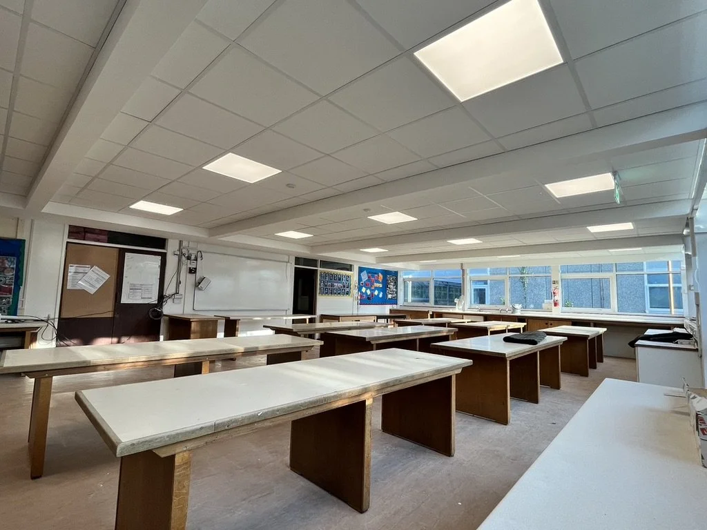Empty classroom with wooden tables, large windows, whiteboards, and overhead lighting.