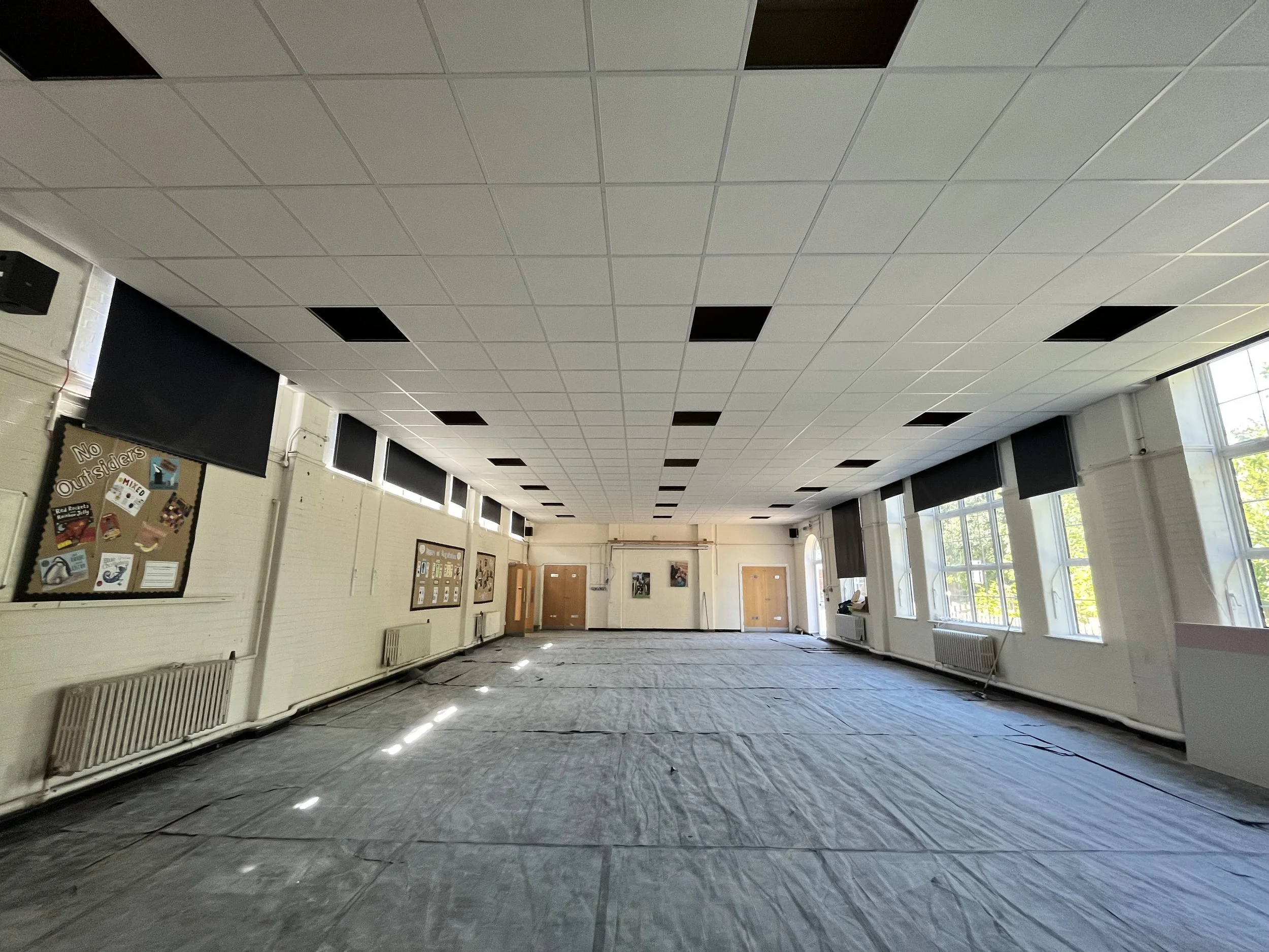 An interior room with a tiled ceiling, large windows on the right side, and chairs and bulletin boards on the left wall, possibly in a school or community center.