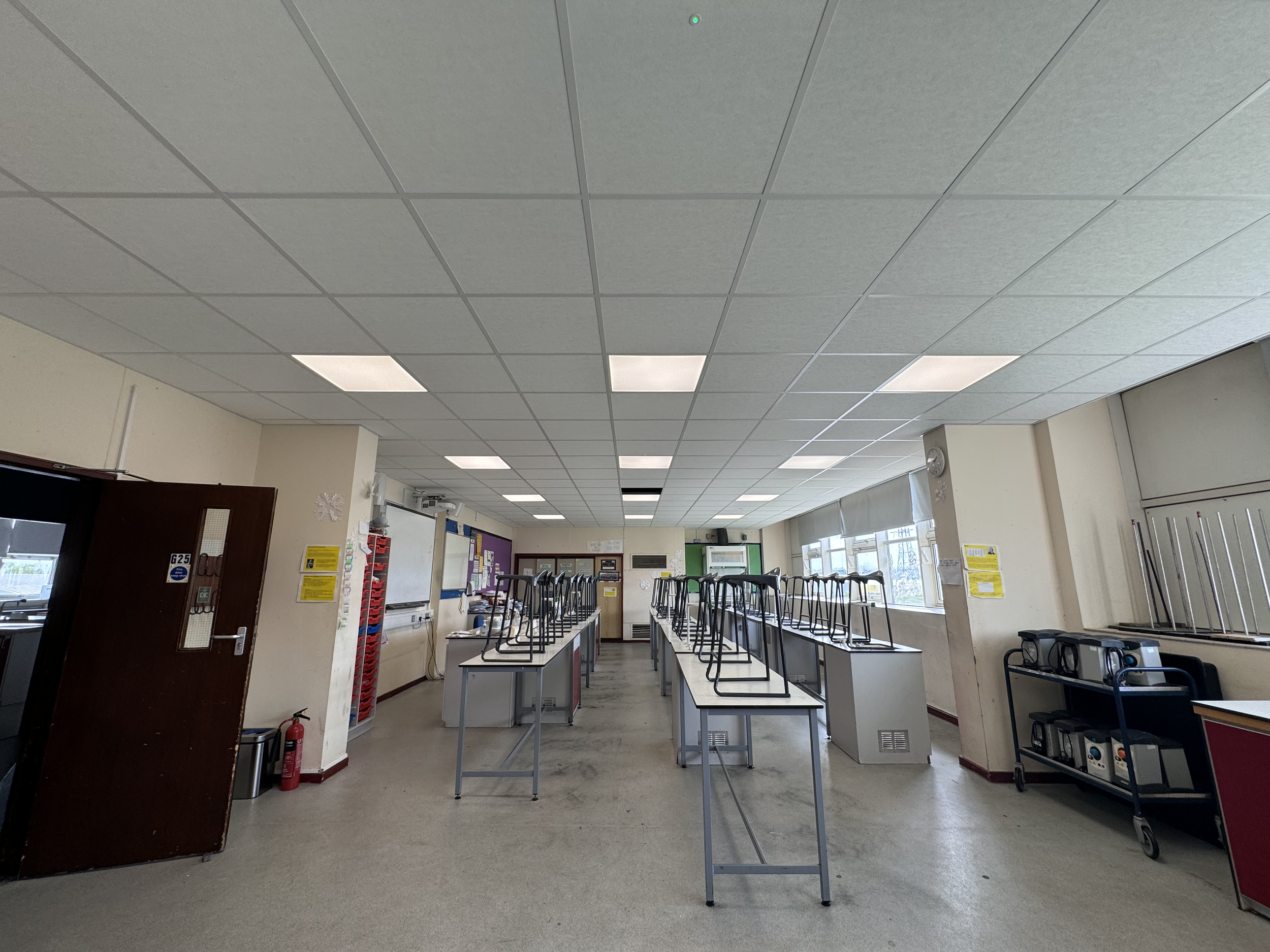 Classroom with ceiling tiles, fluorescent lights, and stacked desks, suggesting it is being set up or cleaned.