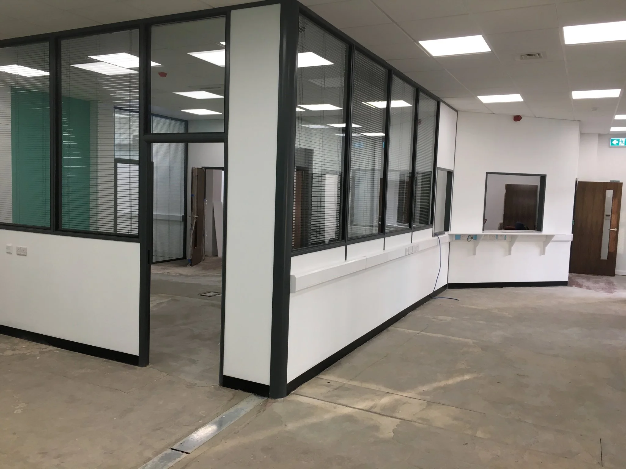 Interior of an office with glass partition walls, ceiling lights, and an emergency exit sign.