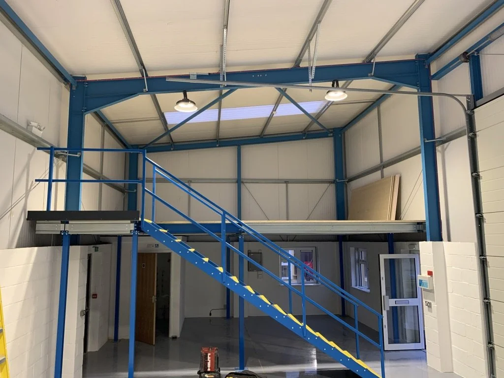 Interior of an industrial warehouse with a blue mezzanine and staircase, wood panels stored on the upper level, and small office rooms on the ground level.