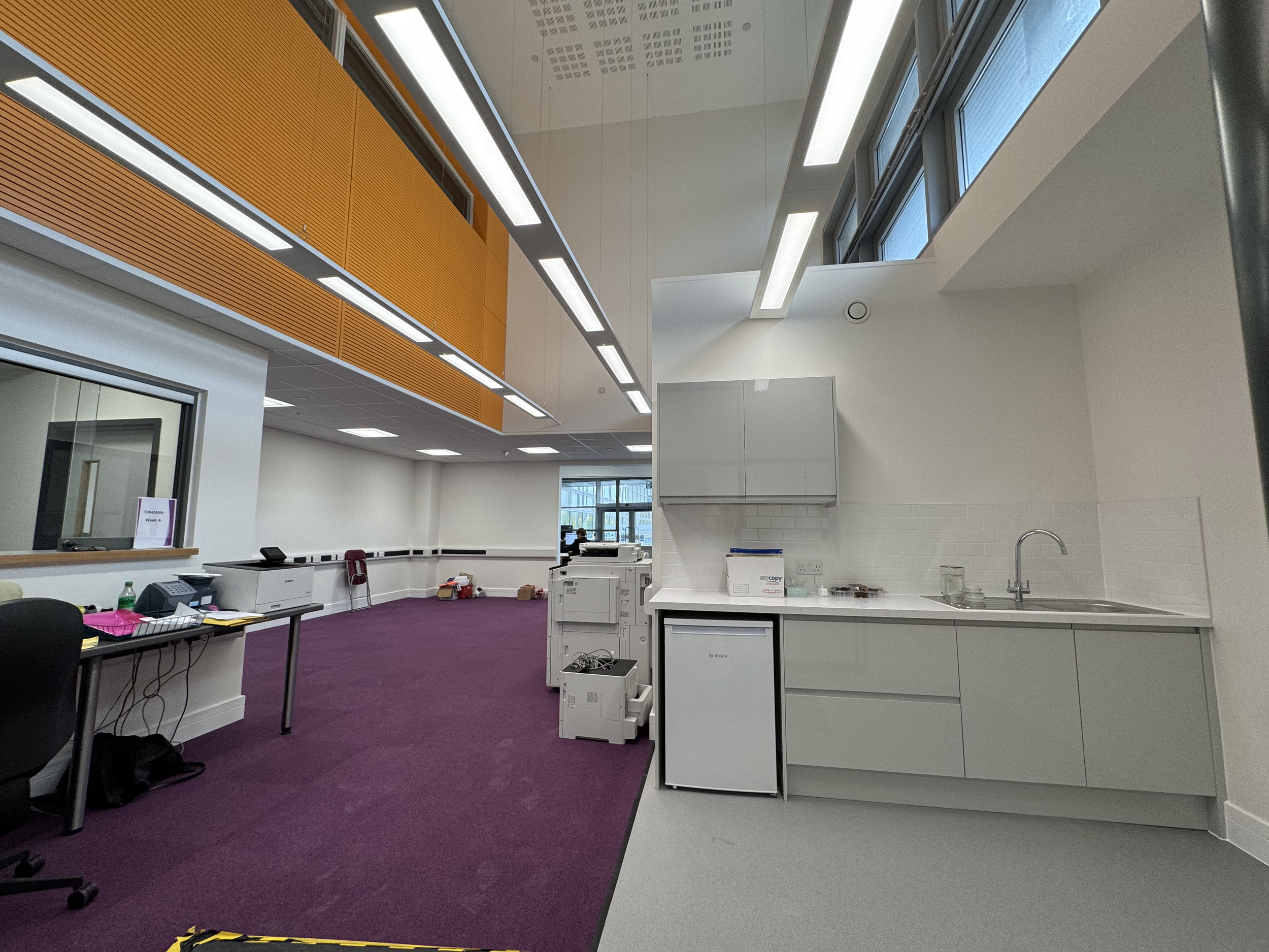 Interior of an office or reception area with white walls, purple carpet, orange wall panels, and a white kitchen counter with a sink and cabinets. There are office supplies, printers, and chairs visible, with two people working in the background.
