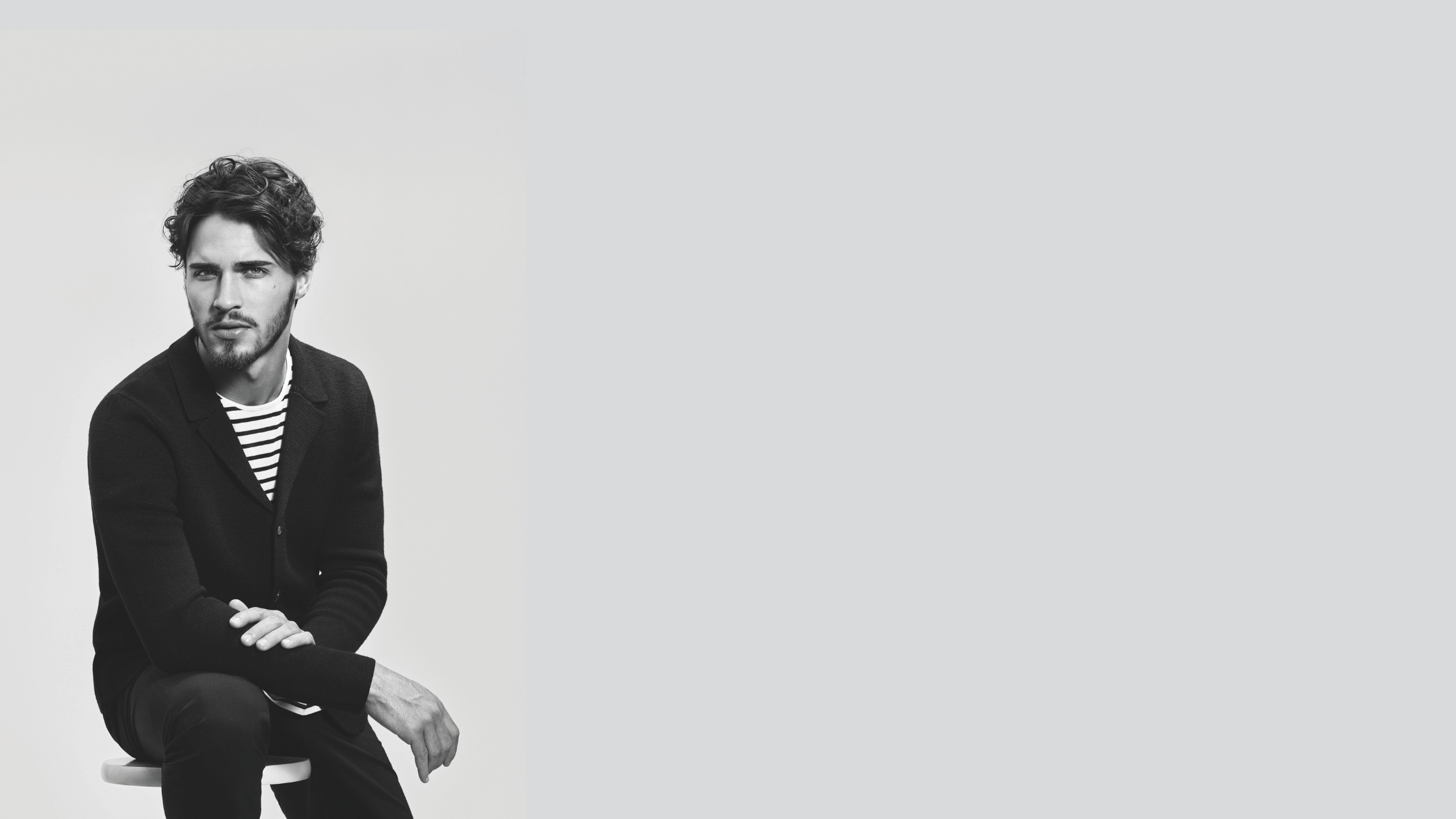 Black and white portrait of a young man sitting on a stool with arms crossed, wearing a dark cardigan and striped shirt, looking at the camera.