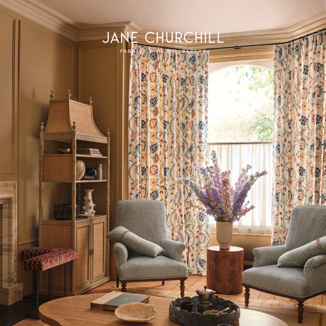 Interior of a cozy living room with two light blue striped armchairs, a coffee table, a vase of purple flowers, patterned curtains, a wooden cabinet, and a window with natural light.