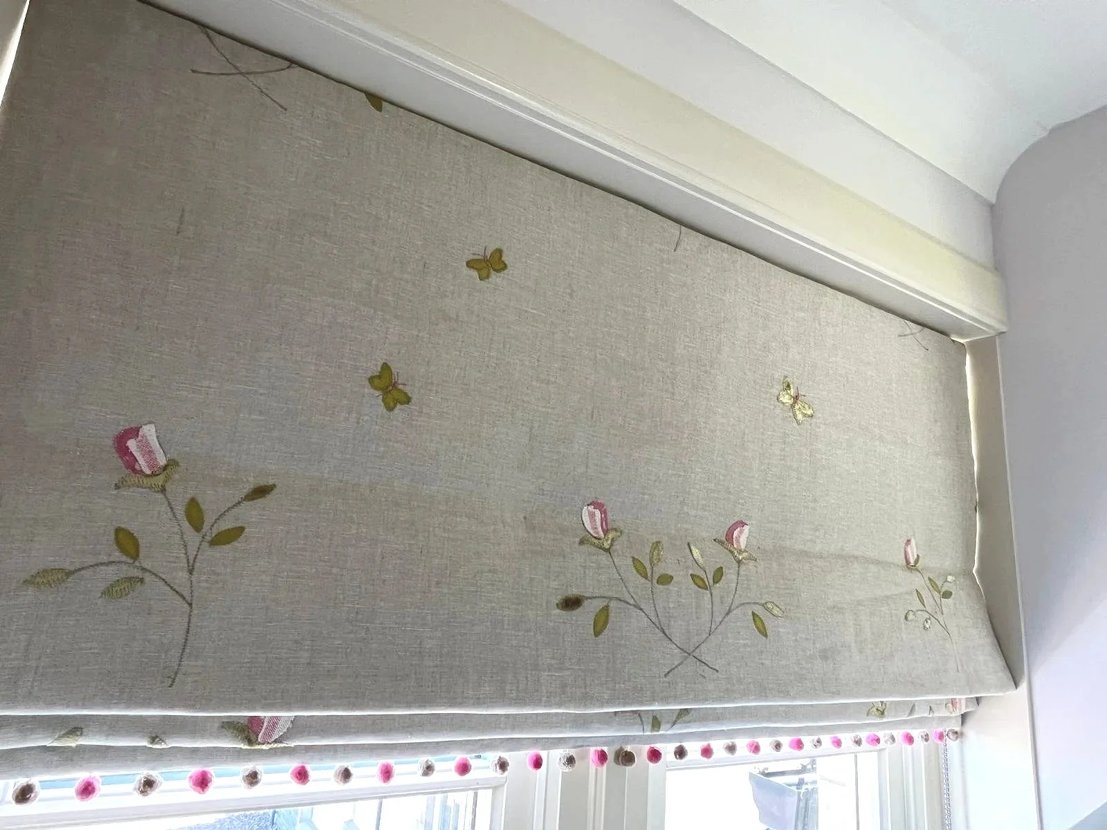 A beige window curtain with embroidered pink roses, green leaves, and yellow butterflies, partially covering a window with pink polka dot trim.