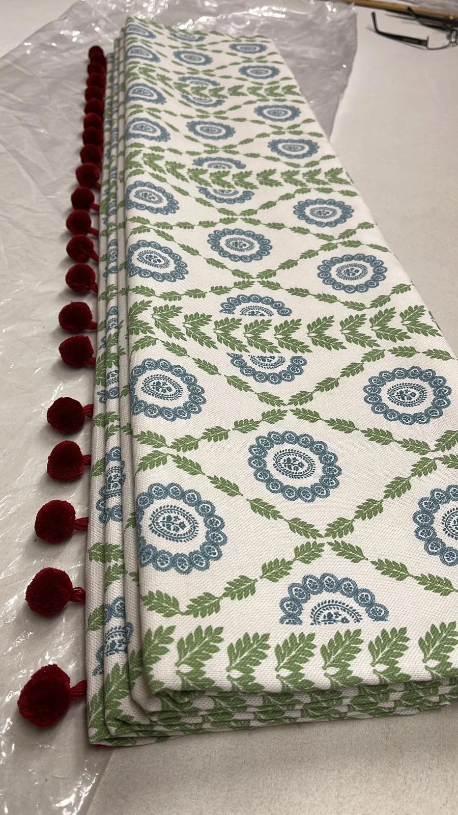 Folded tablecloth with a blue and green floral pattern and red pom-pom trim, placed on a table covered with plastic.