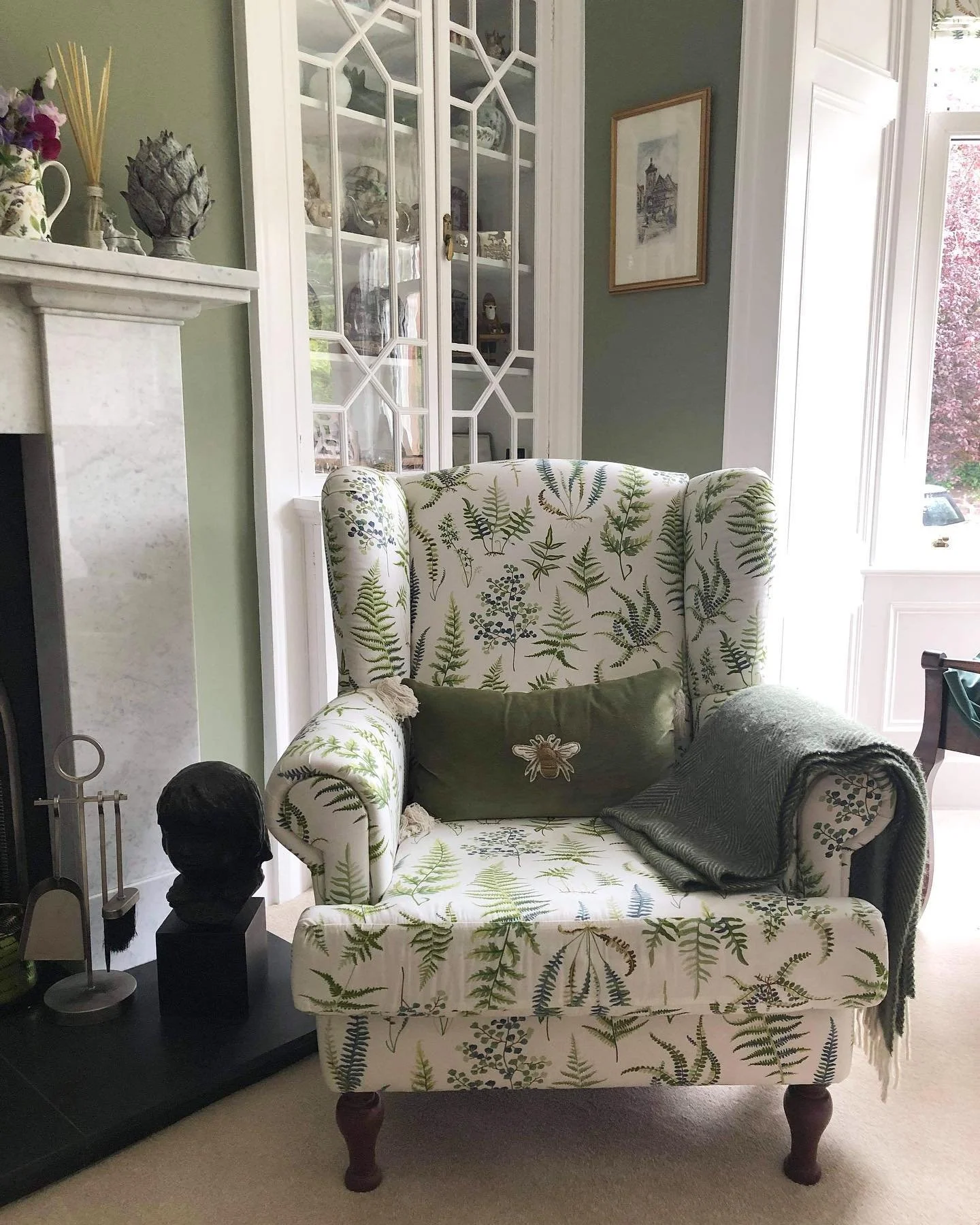 A floral-patterned armchair with a green pillow and a gray throw blanket in a cozy living room. There's a white decorated fireplace, a glass-fronted cabinet, and a framed picture on the wall. Sunlight streams through a large window outside.