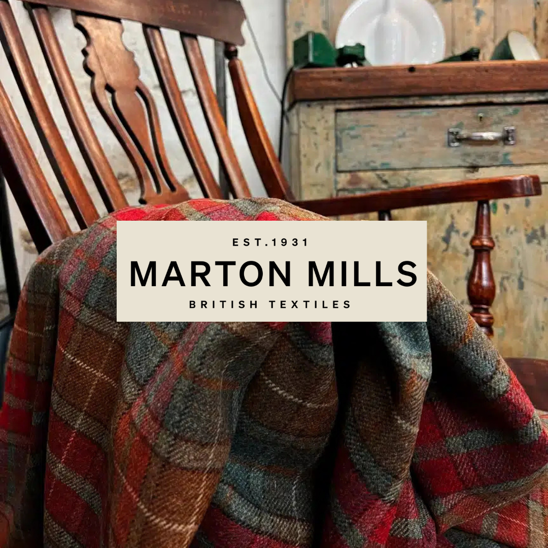 A wooden chair with a plaid fabric draped over it, positioned in a rustic setting with a distressed wooden dresser and dishes in the background, featuring a logo for Marton Mills, British textiles, established 1931.