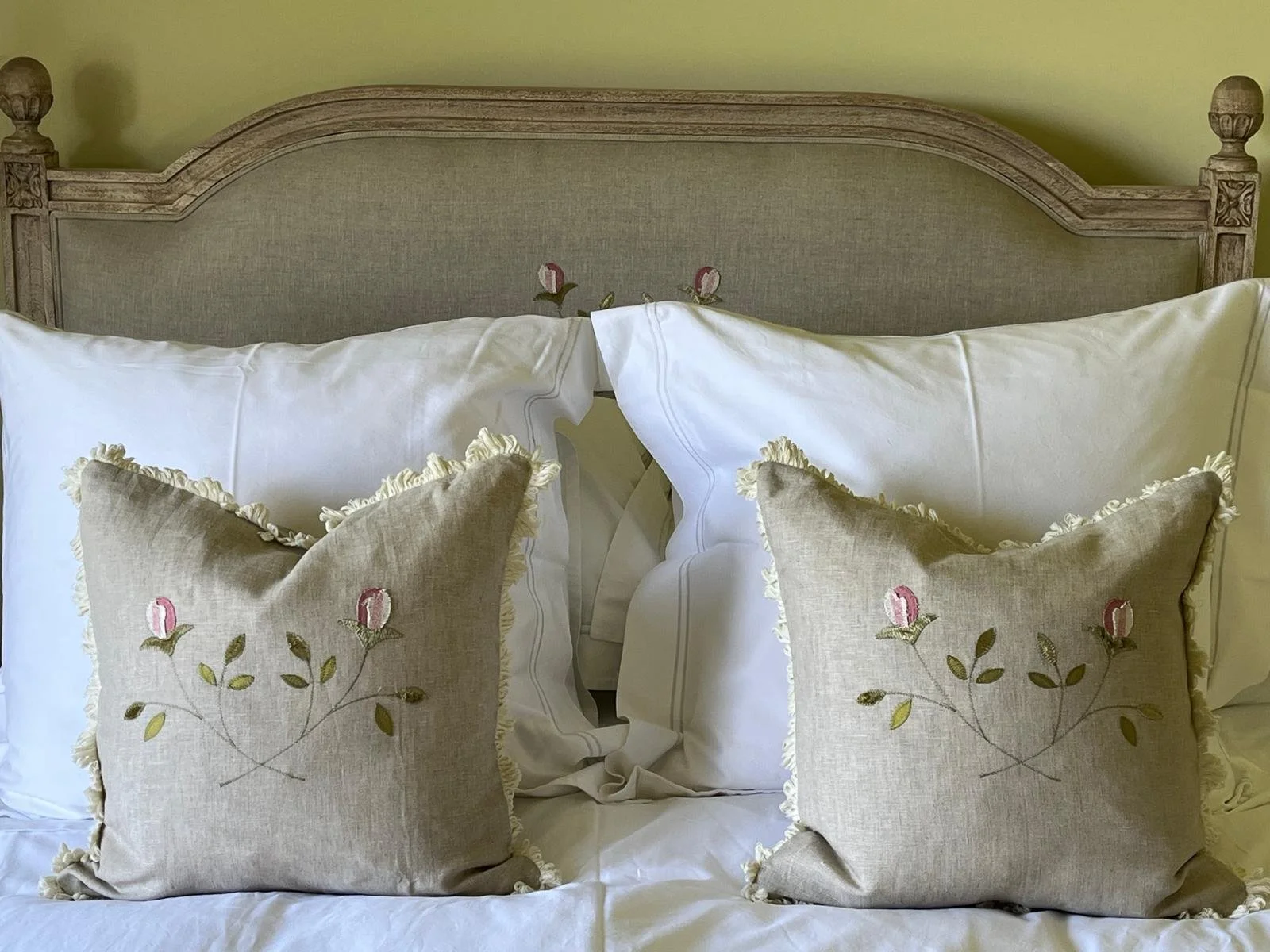 A bed with a wooden headboard, two large white pillows, and two decorative beige pillows with embroidery of pink roses and green leaves, fringed edges, on white sheets.