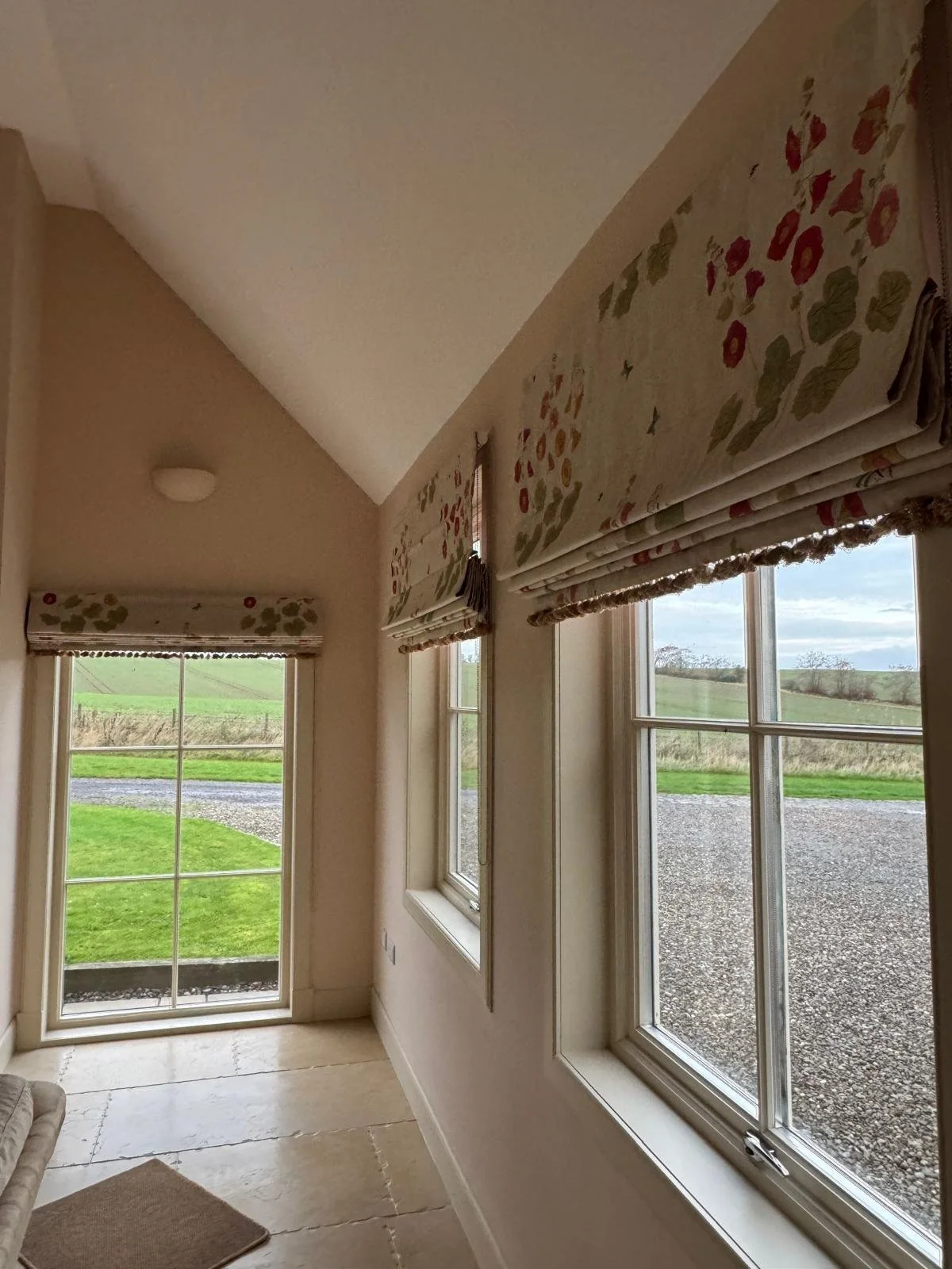 A sunlit room with three windows and a glass door, decorated with floral window coverings, overlooking a green grassy landscape with a gravel driveway.