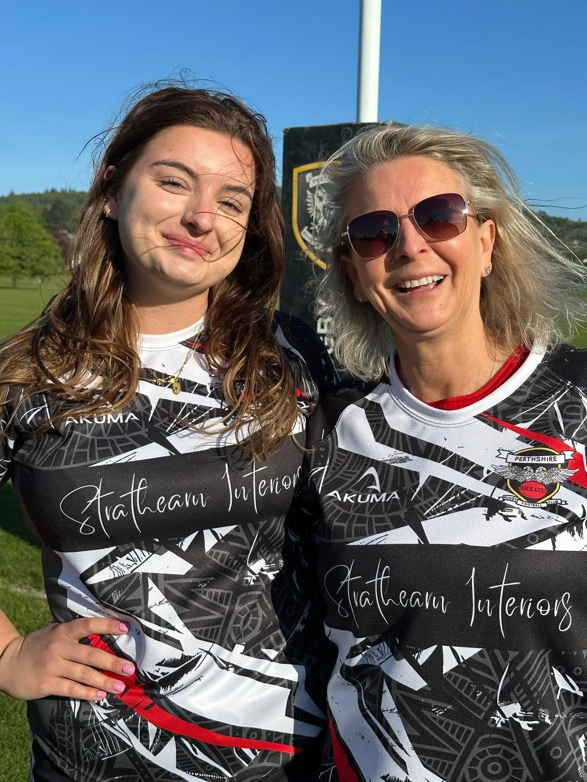 Two women wearing black and white sports jerseys with red accents, standing outdoors with a sunny sky, a field, and a shield-shaped sign in the background. One woman has sunglasses and blond hair, the other has brown hair and is smiling.