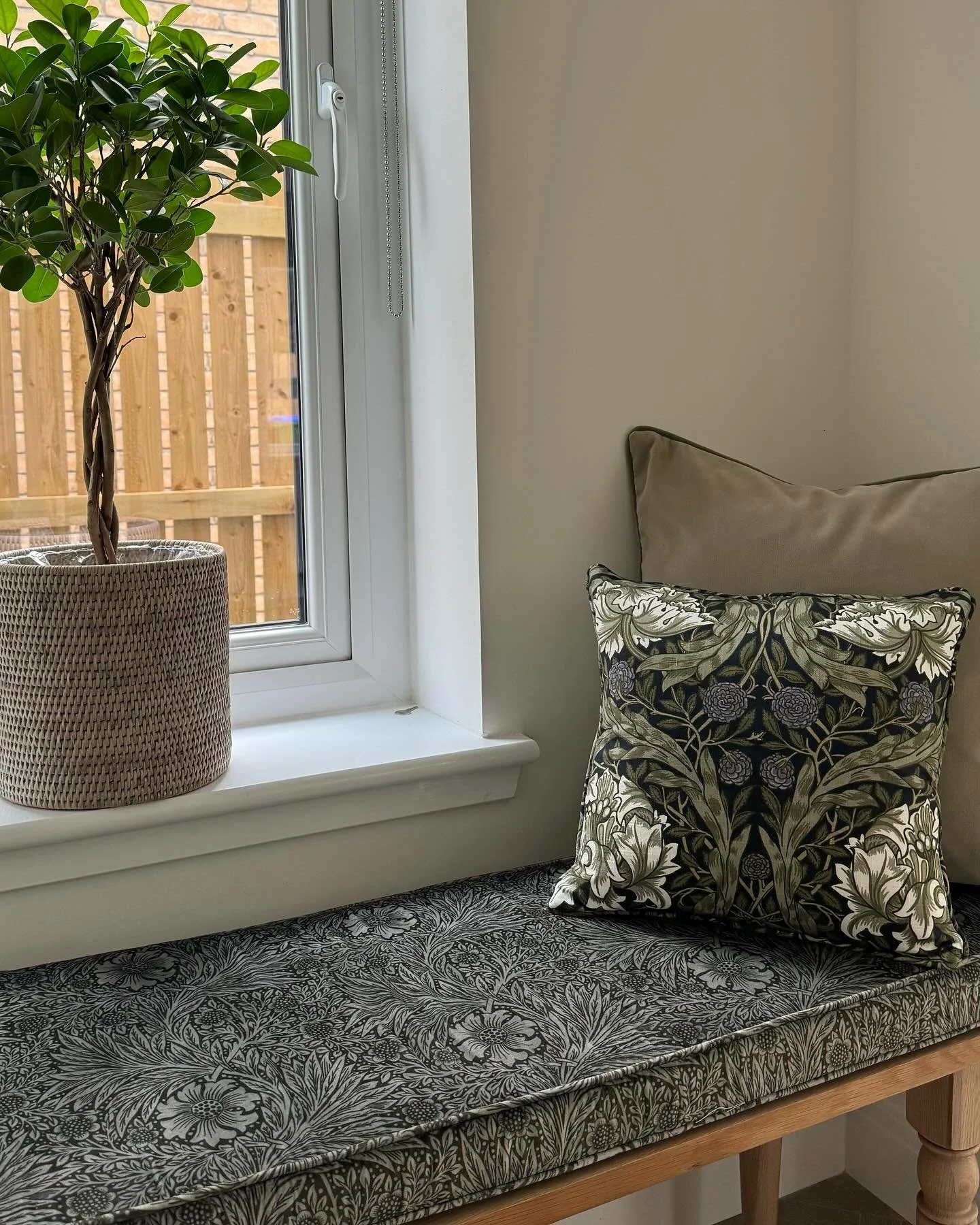 Window with a potted plant on the sill and two decorative pillows on a cushioned bench indoors. with a custom soft furnishing window seat