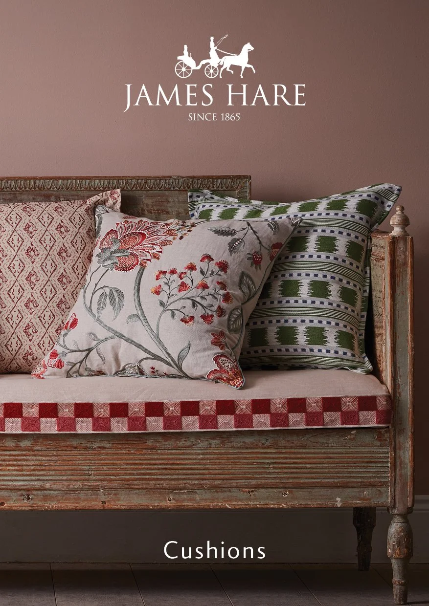 A rustic bed with a wooden frame, decorated with various patterned cushions and pillows. The wall behind has a sign that reads 'James Hare Since 1865' with a vintage horse-drawn carriage logo. A caption at the bottom says 'Cushions'.