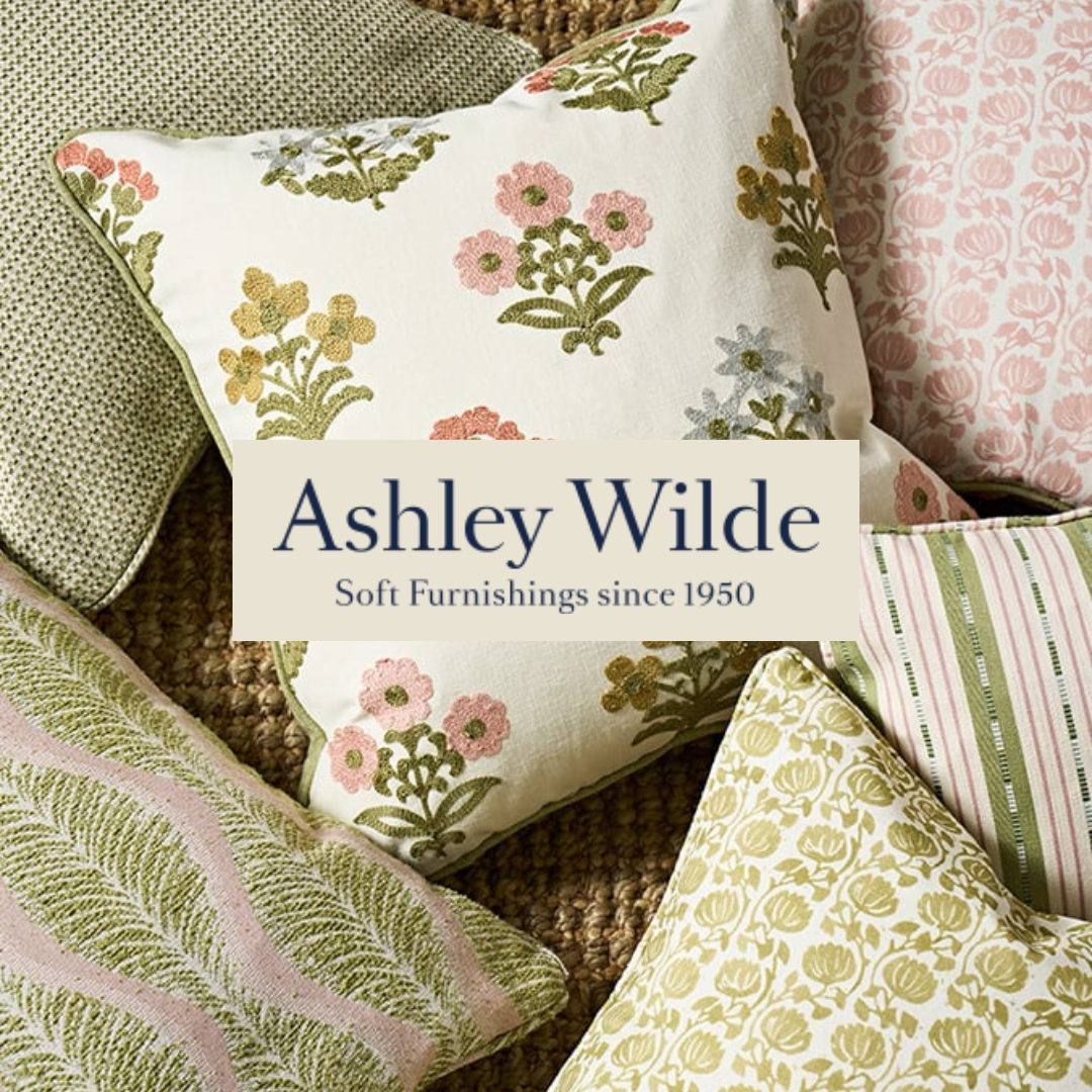 Collection of decorative pillows with floral and striped patterns, over a textured brown surface, featuring the Ashley Wilde logo and text for a home furnishings brand.
