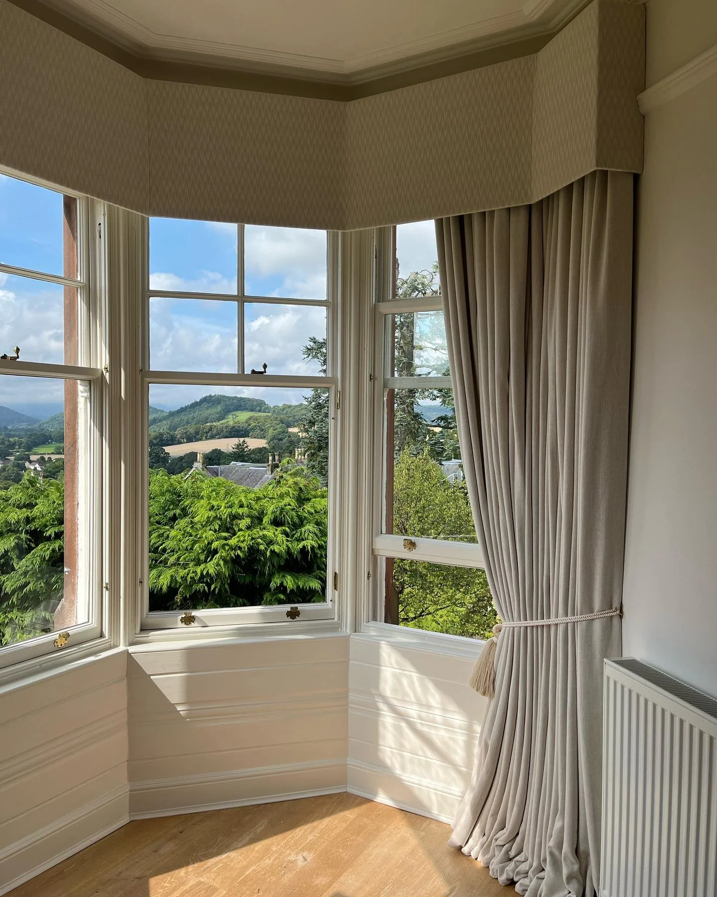 Room with large bay window showing a view of green trees, hills, and cloudy sky, with white curtains pulled to one side.