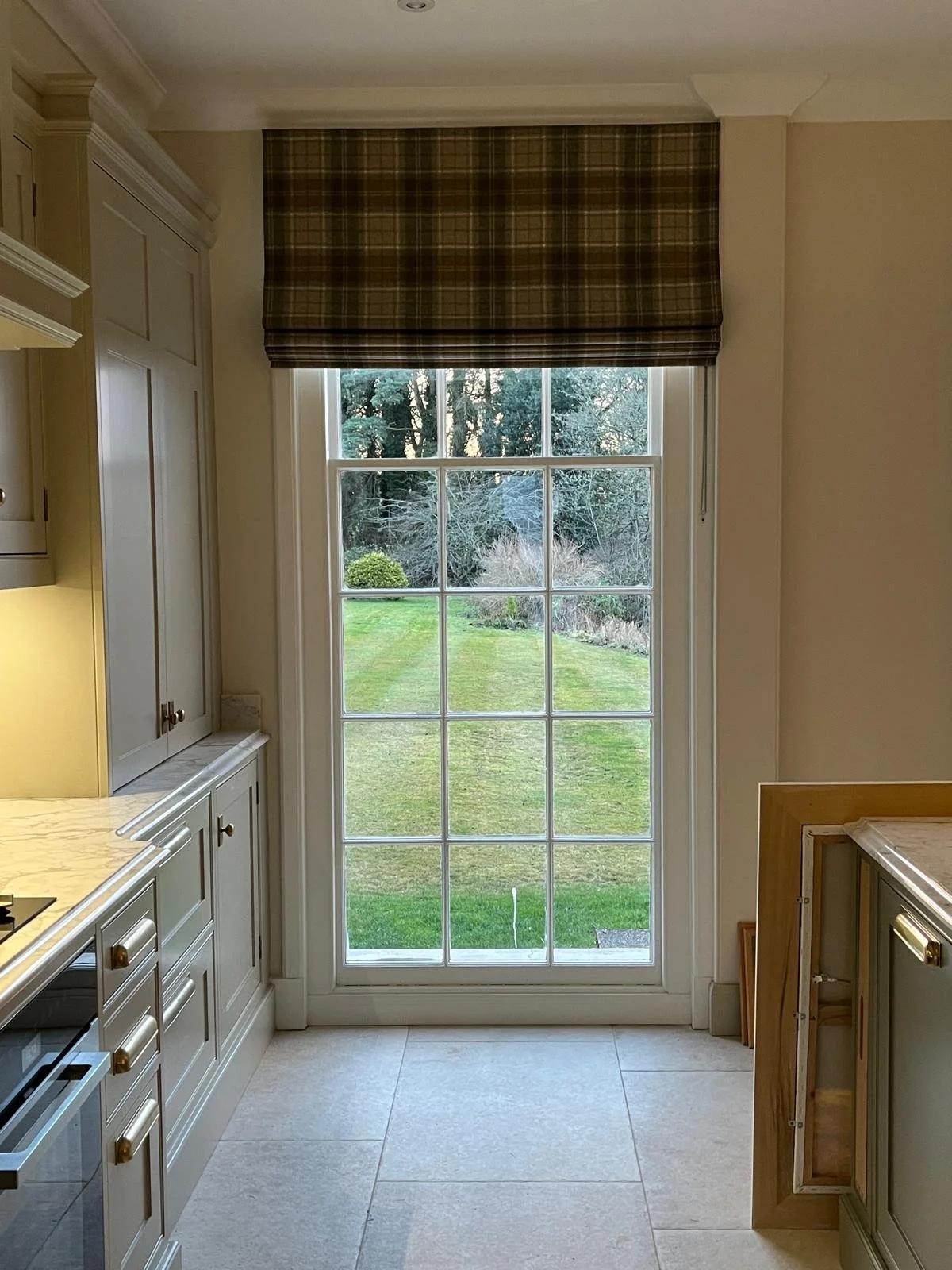 Made-to-measure Moon tweed Roman blind in a classic check pattern, fitted in a bright kitchen window with garden view, providing soft light control and timeless style. In perthshire