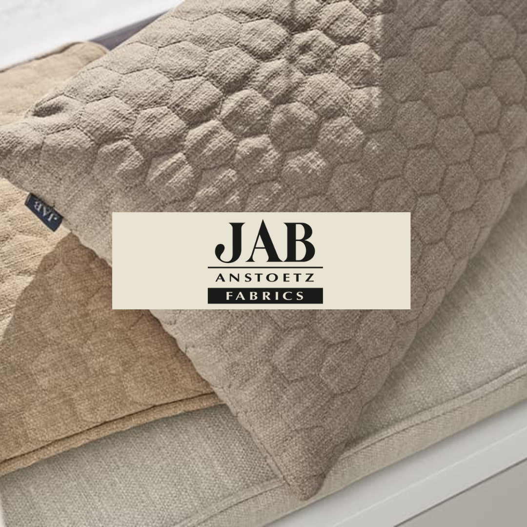 Close-up of three fabric cushions in beige tones, with a label on the left cushion, and a logo overlay that reads "JAB ANSTOETZ FABRICS".