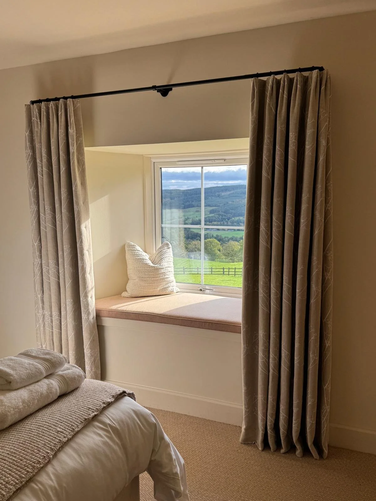 A cozy bedroom with a window seat overlooking a lush, green landscape with rolling hills and trees, framed by bespoke beige curtains.
