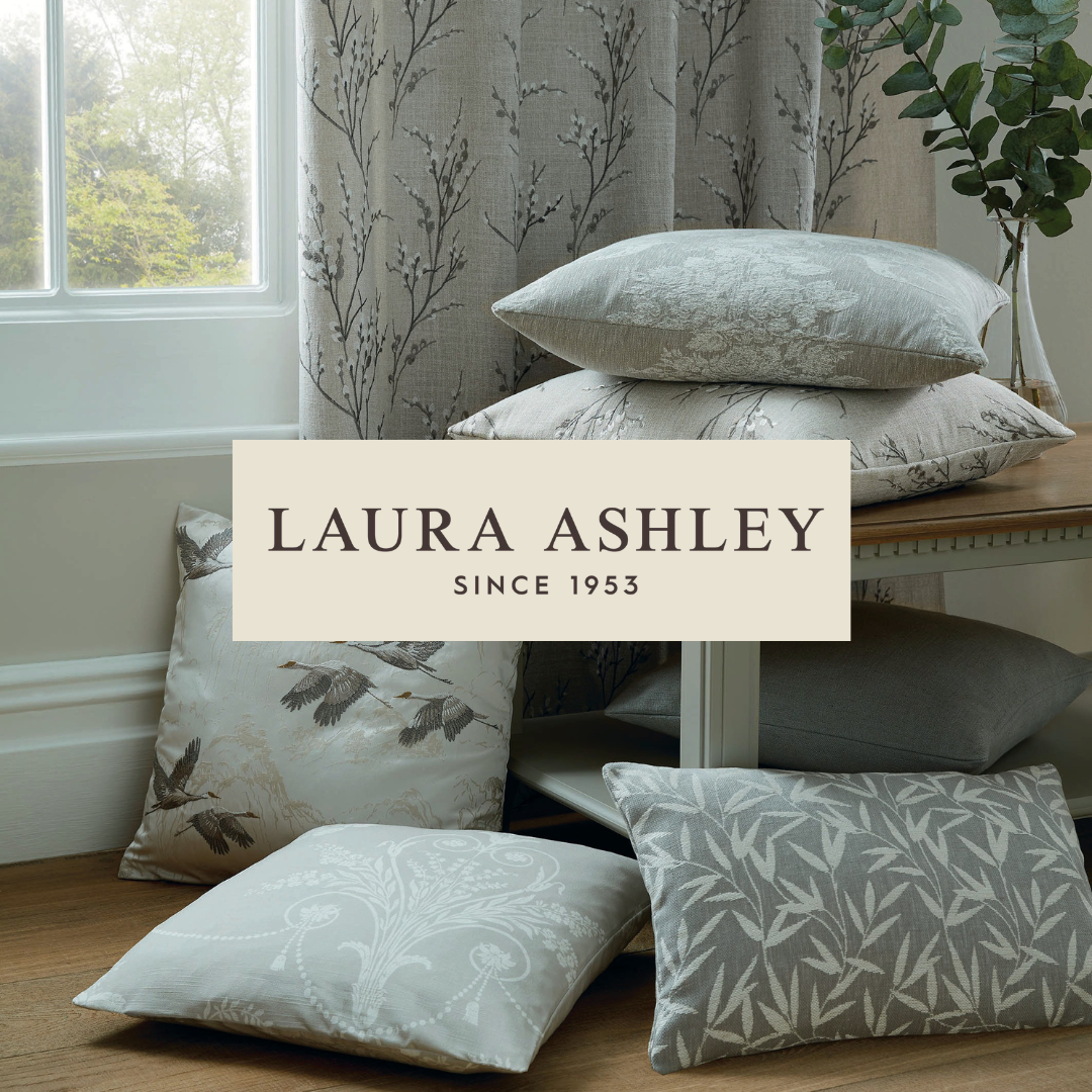 Stacked and scattered decorative pillows with floral and bird patterns in front of a window with tree view, curtains, and a side table with greenery.