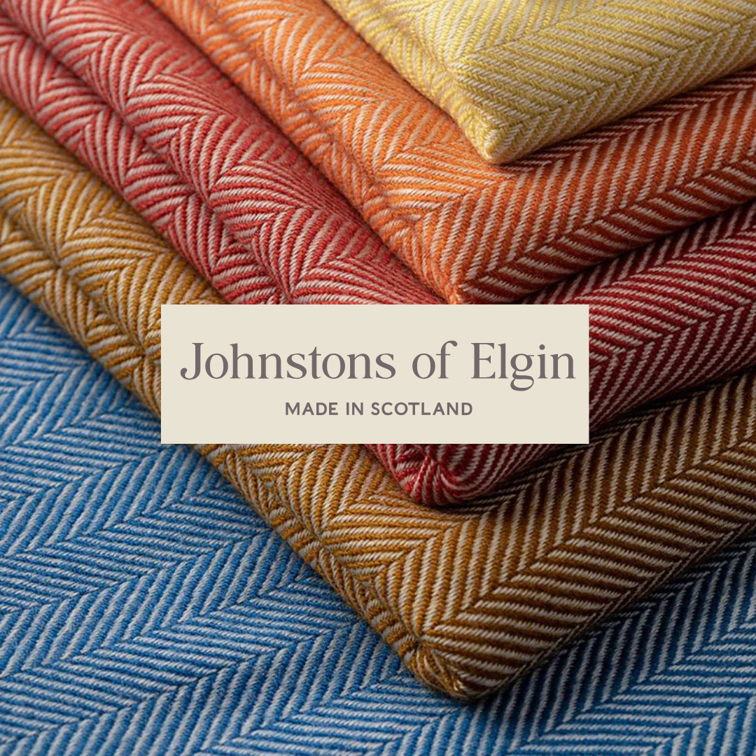 Stack of folded herringbone-patterned fabric in various colors including red, orange, yellow, beige, and blue, with a label that reads 'Johnstons of Elgin, Made in Scotland'.