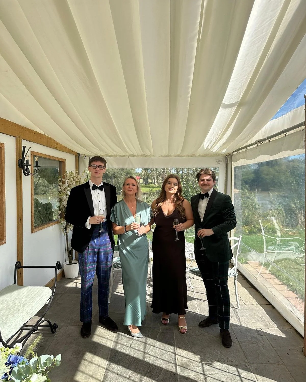 Four people dressed in formal attire standing inside a white tent with draped ceiling, holding glasses of champagne and posing for a photo.
