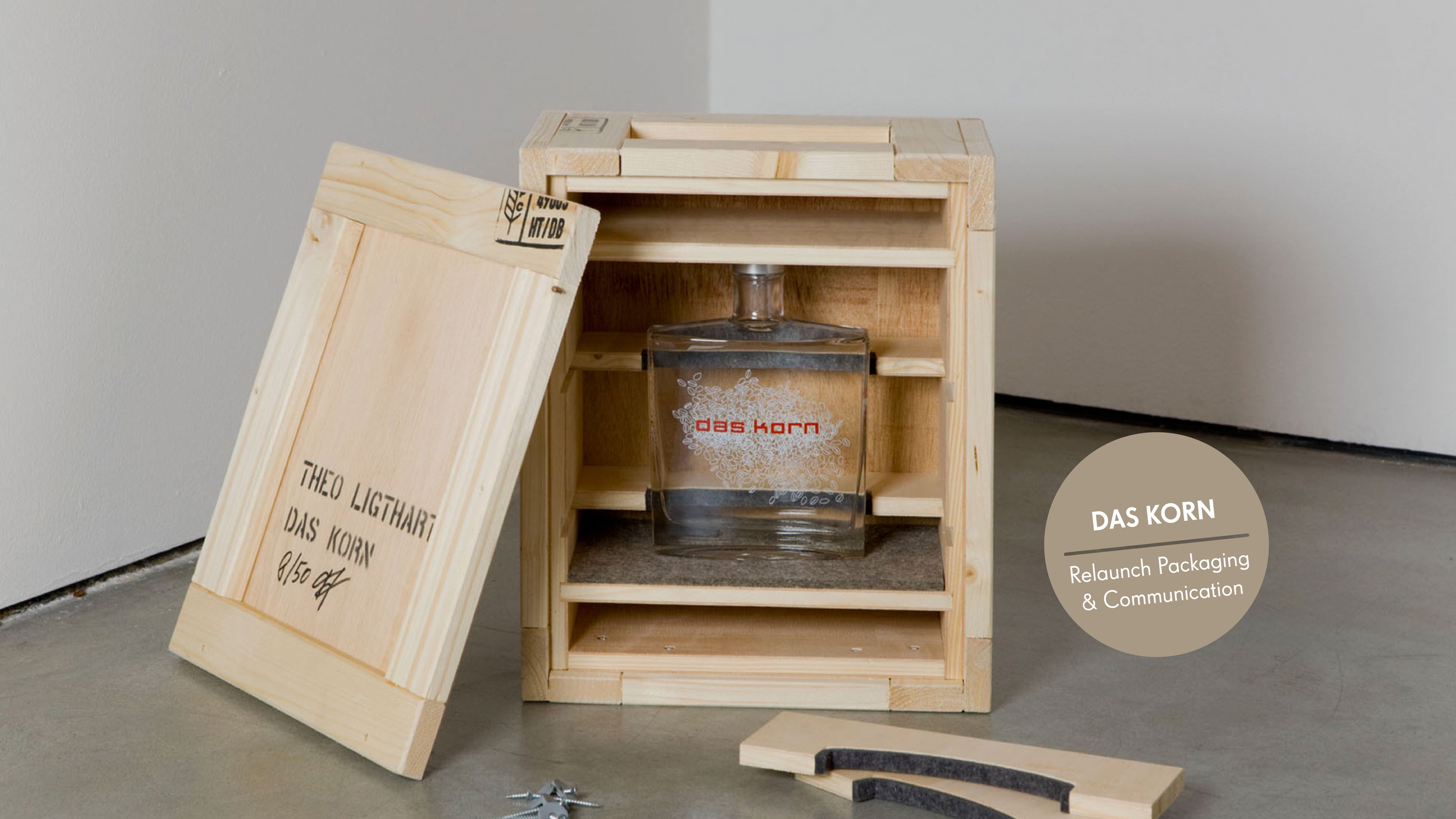 Holzkiste mit offenem Deckel und eine Glasflasche mit Etikett, das 'das Korn' liest, steht vor einer weißen Wand. Es liegen Werkzeuge und Holzstücke auf dem Boden.