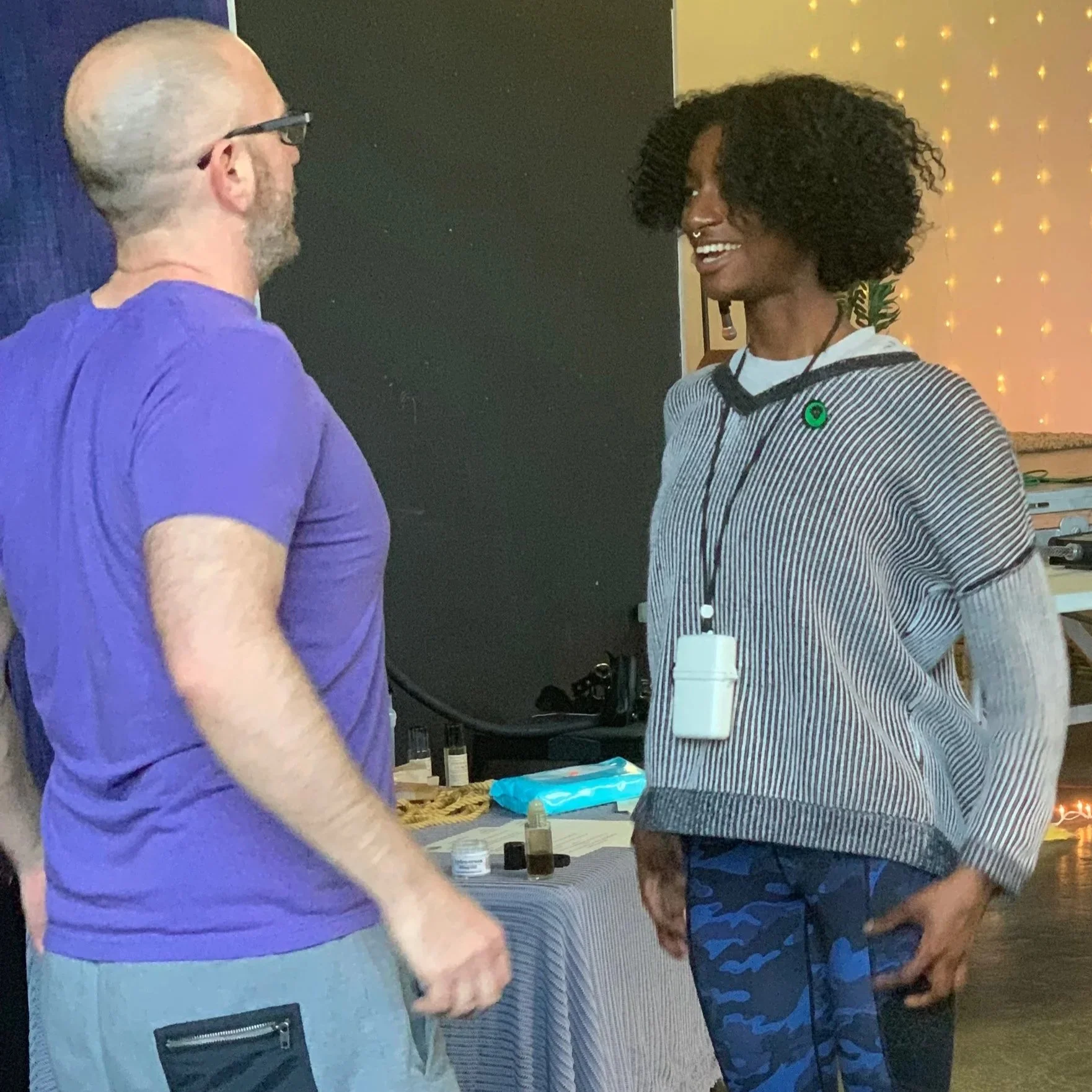 A man with glasses and a beard wearing a purple t-shirt talking to a woman with curly hair, wearing a striped sweater and blue camouflage pants, in a room with warm lighting and decorative string lights.