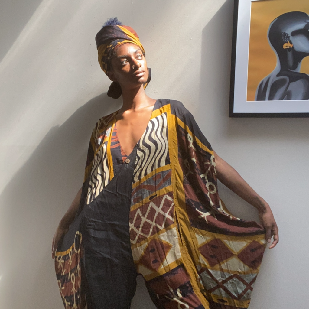 A woman with dark skin and purple hair covered with a colorful headwrap, standing against a plain gray wall. She is wearing a loose, patterned kimono-style cover-up with various earthy tones and geometric designs.