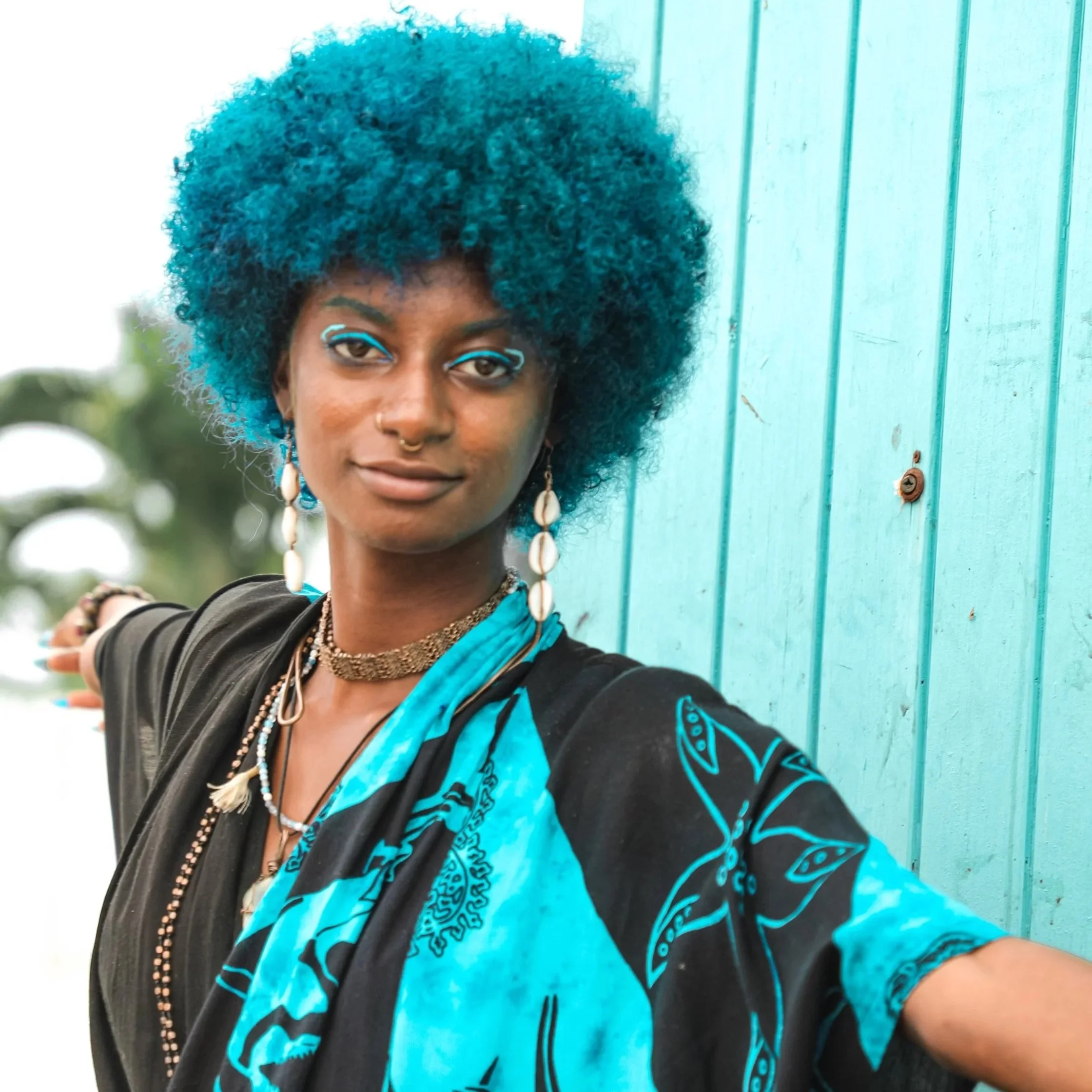 A woman with dark skin and curly blue hair wearing a blue and black patterned shawl, with earrings, necklaces, and face makeup, standing next to a turquoise wall with a tree in the background.