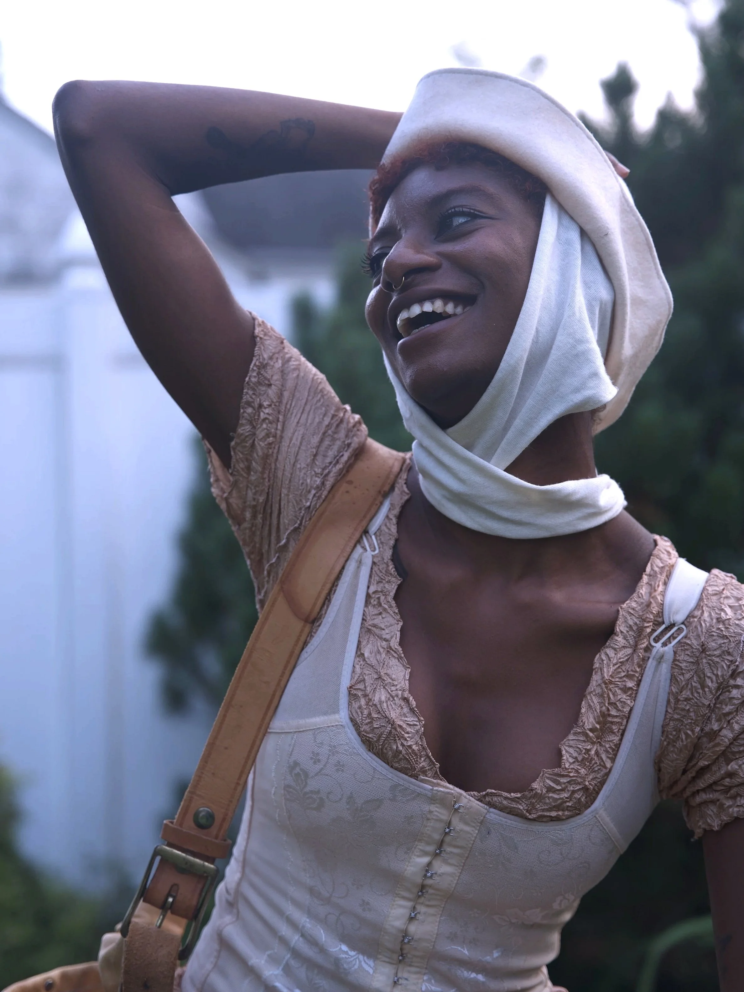 Smiling Indigo Davis with short hair and septum piercing, wearing a cream-colored vintage dress with lace and button details, head wrapped in a white cloth, outdoors in a garden.