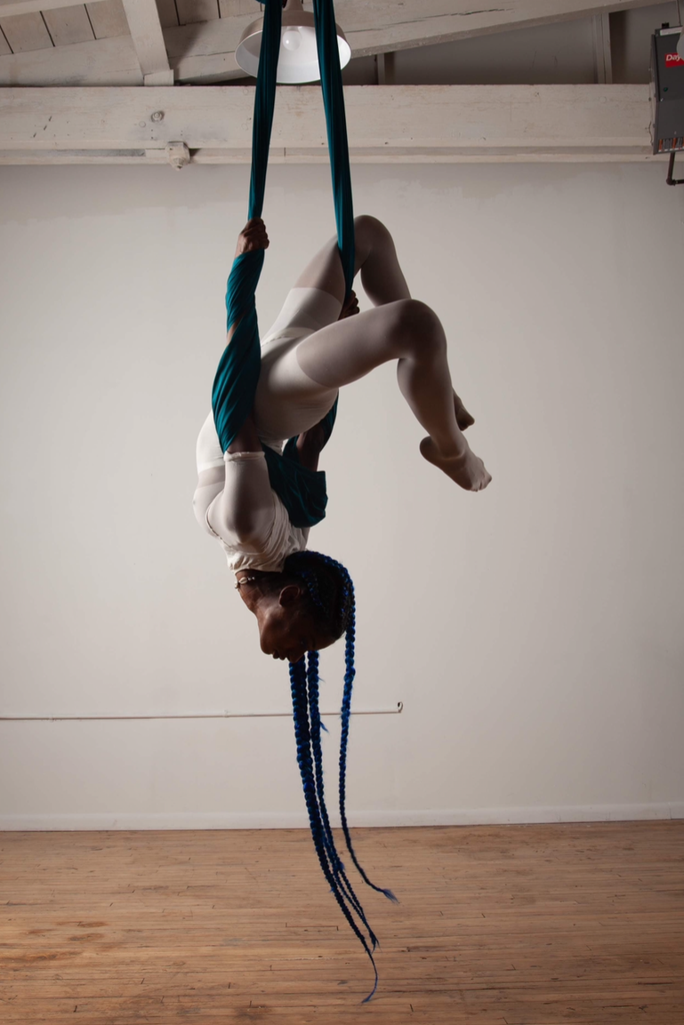 A person with long blue braids performing an aerial silk routine upside down in a studio with white walls and wooden floors.