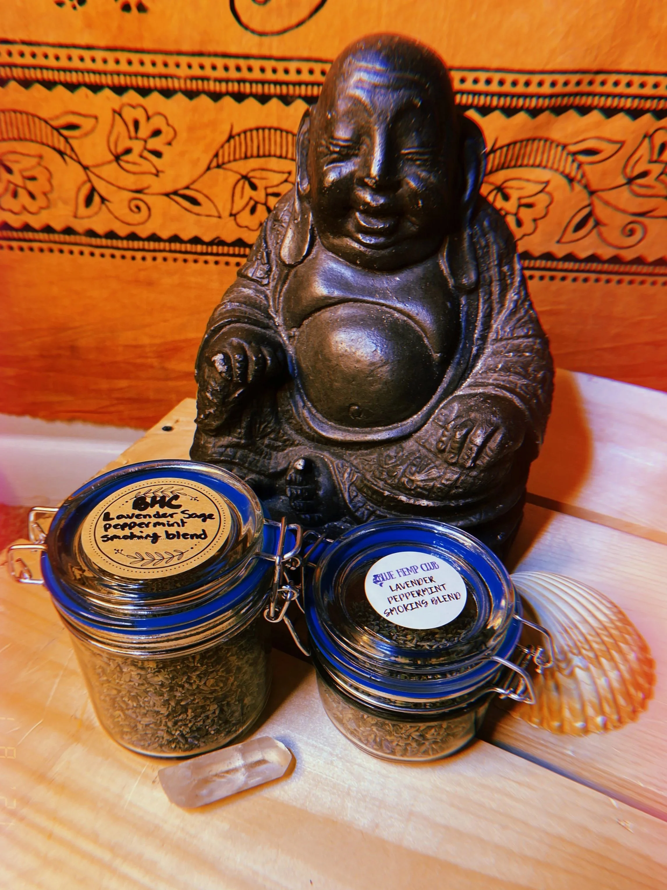 A Buddha statue is placed on a wooden surface behind two small glass jars labeled 'Lavender Sage Pepperment Smokin' Blend' and 'Lavender Pepperment Smokin' Blend.' An orange shell is also on the surface. The background features a decorative, patterned tapestry.