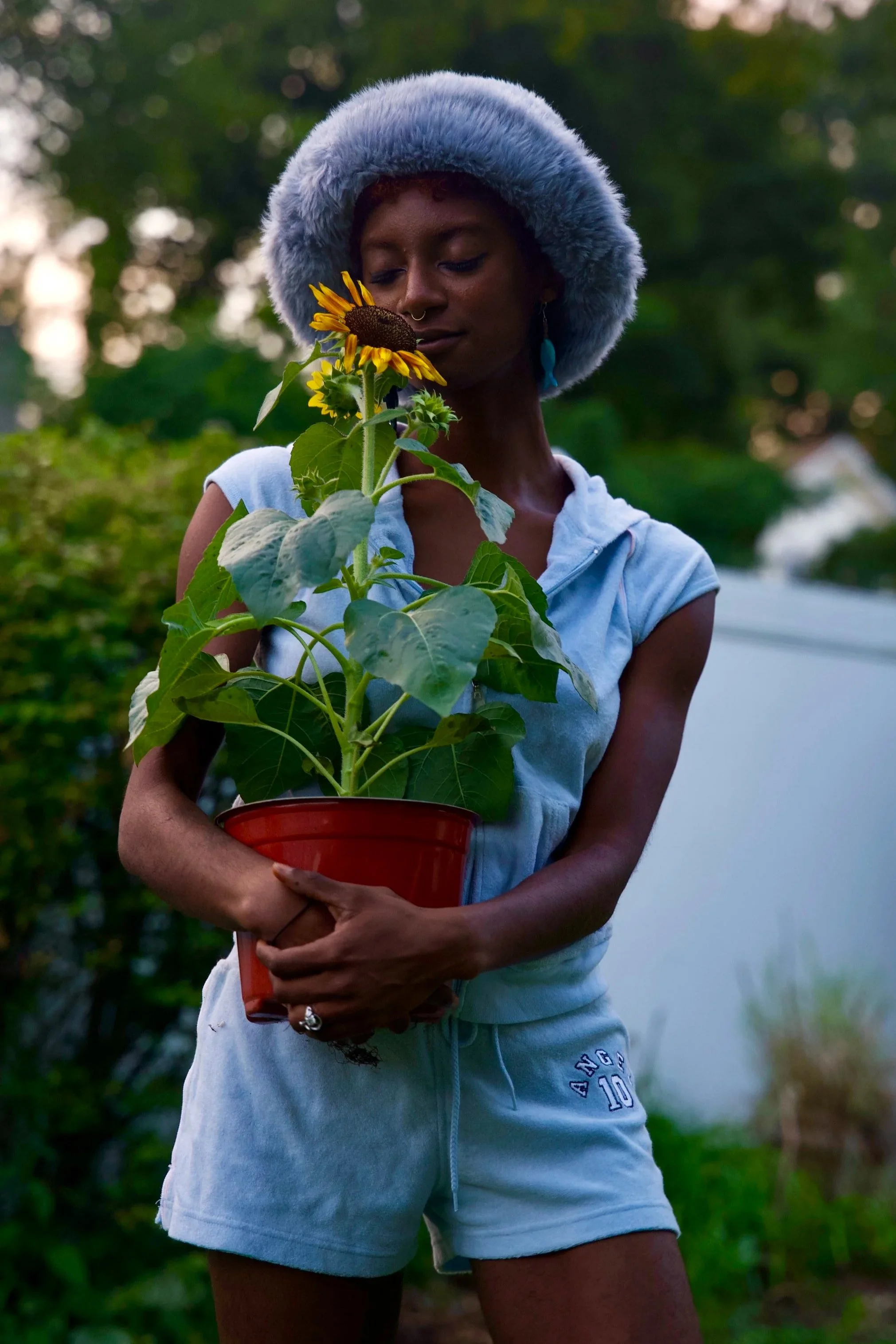 Woman with dark skin, wearing a blue furry hat and light blue hoodie, holding a red pot with a sunflower plant, outdoors during evening.