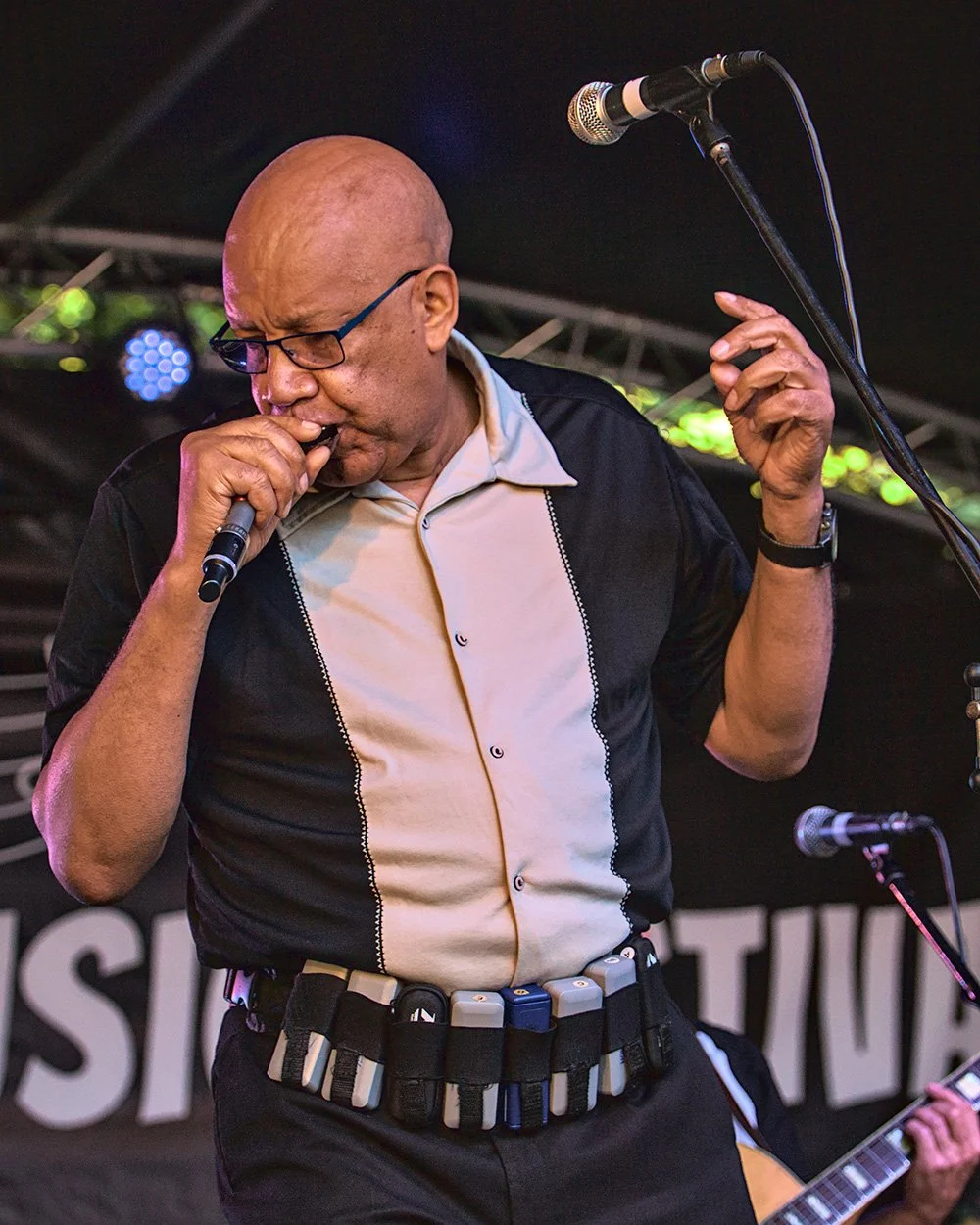 A bald man wearing glasses, a retro black and white shirt, and a belt with various items attached, is singing into a microphone on stage while holding another microphone in his hand. He appears emotional with his eyes closed and head tilted down. A person playing guitar is partially visible in the background.
