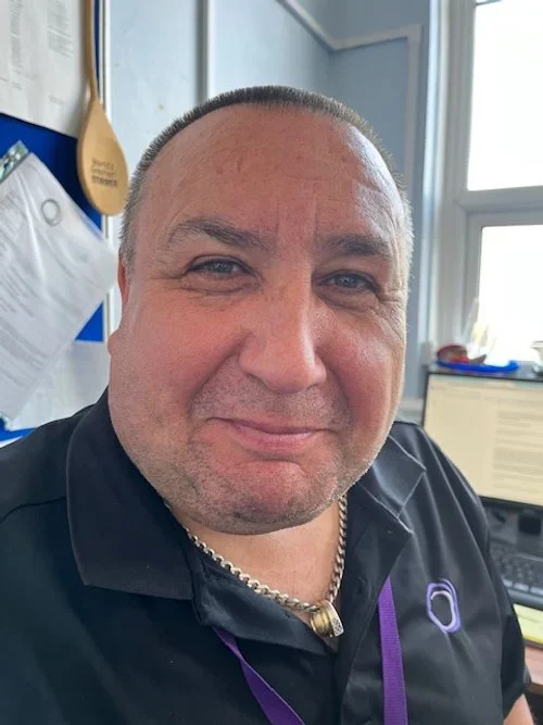 A man taking a selfie in an office with a computer, papers, and a window in the background.
