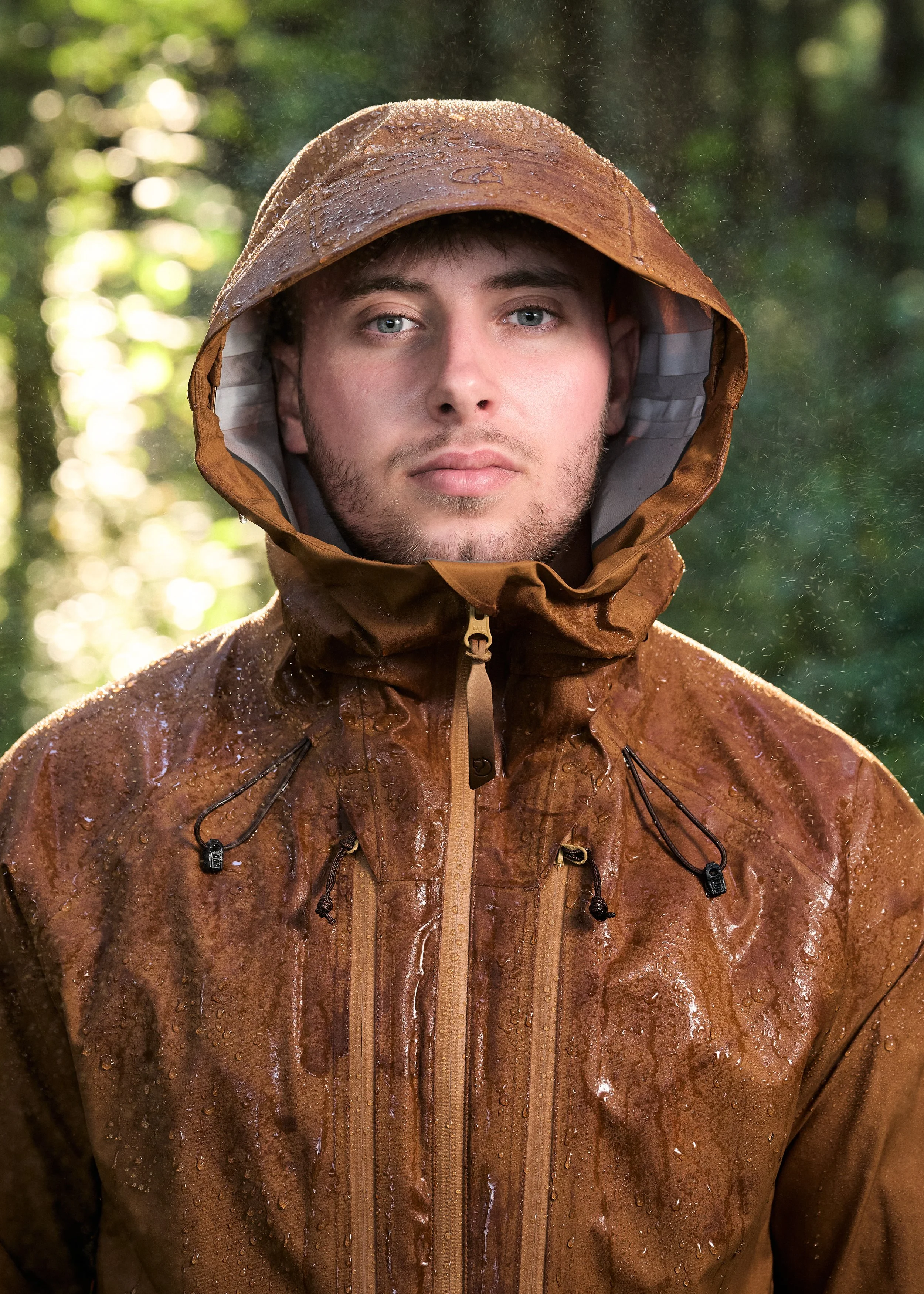 Young man wearing a brown waterproof hooded jacket with raindrops on it, standing outdoors in a wooded area.