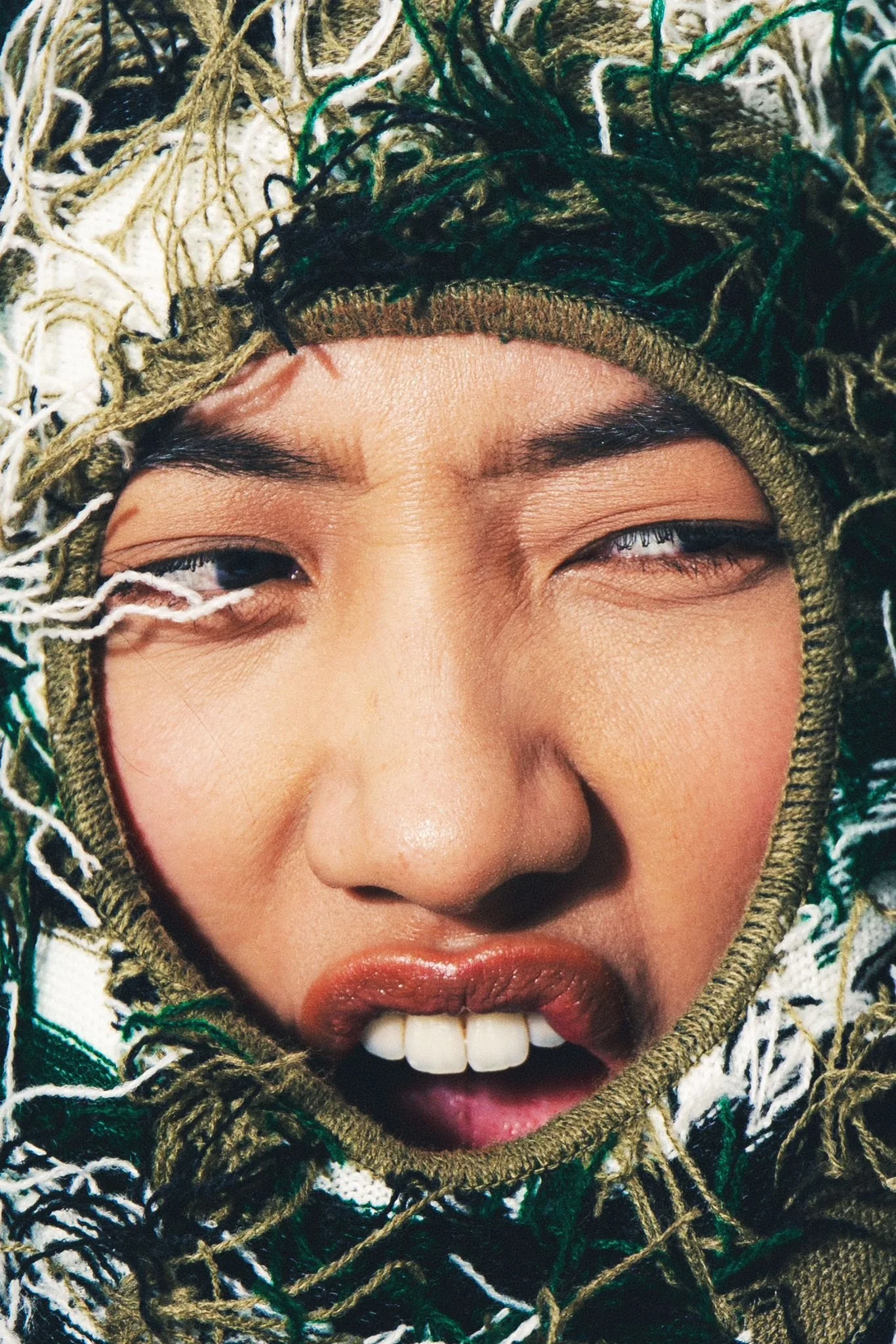 Close-up of a person's face with one eye closed and the other slightly open, partially covered by a woven scarf with fringes, highlighting their eyebrows, nose, and lips.