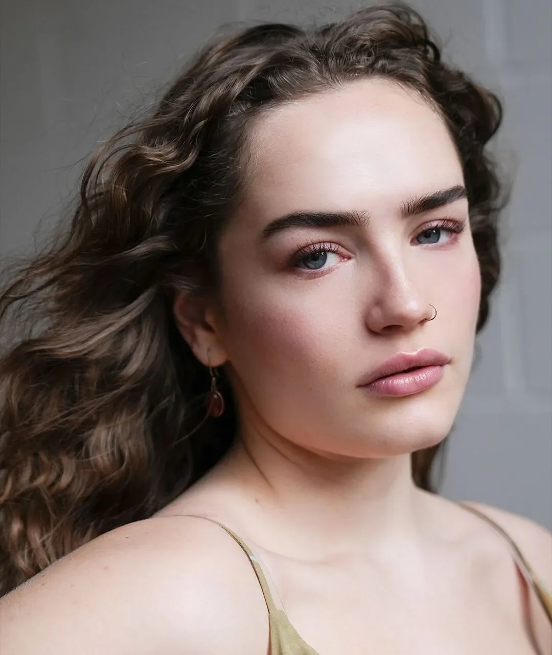 A young woman with light skin, wavy brown hair, blue eyes, and a nose ring, looking at the camera with a neutral expression.