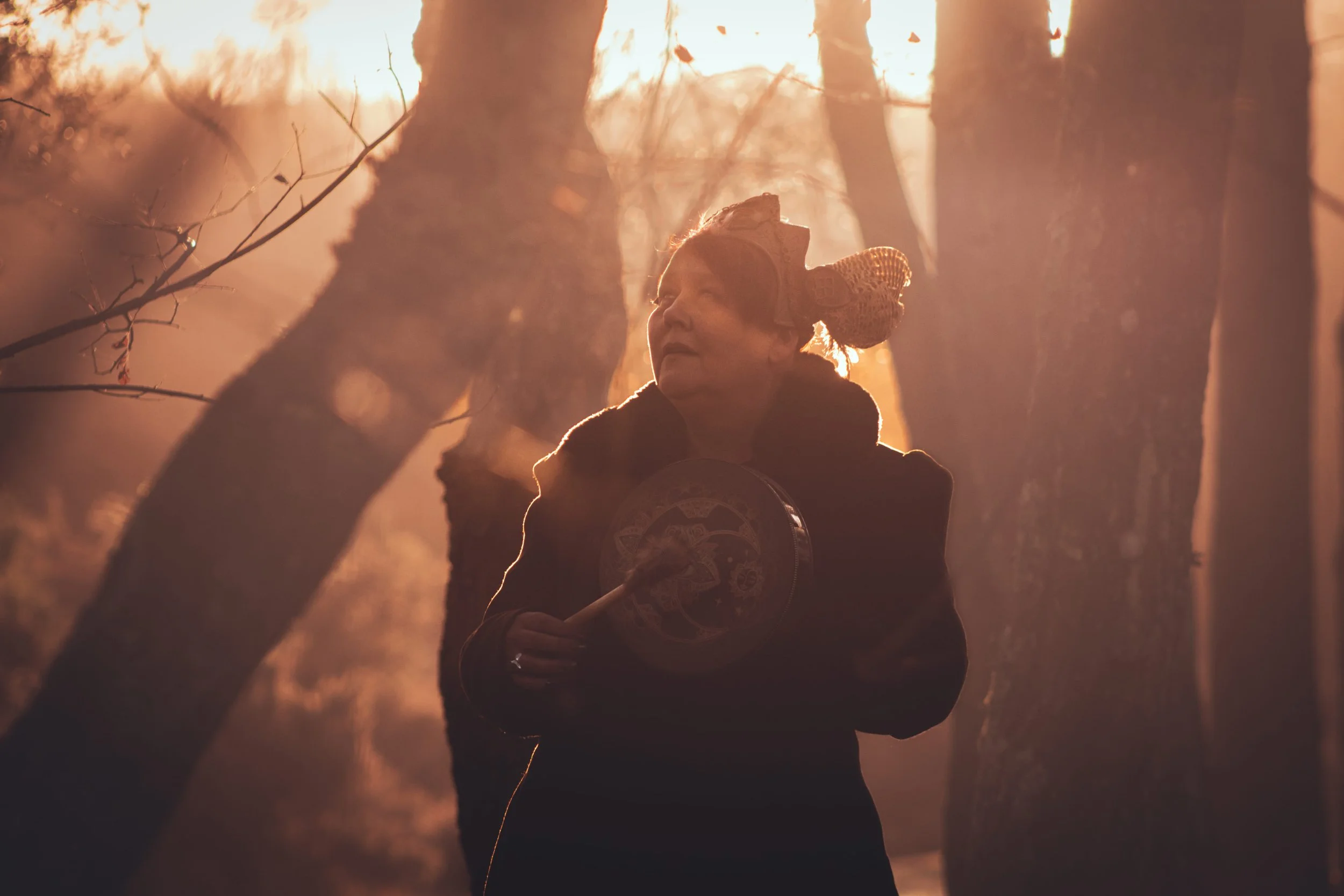 An elderly woman with traditional Asian attire holding a fan, walking among trees in a forest during sunset, backlit with warm light.