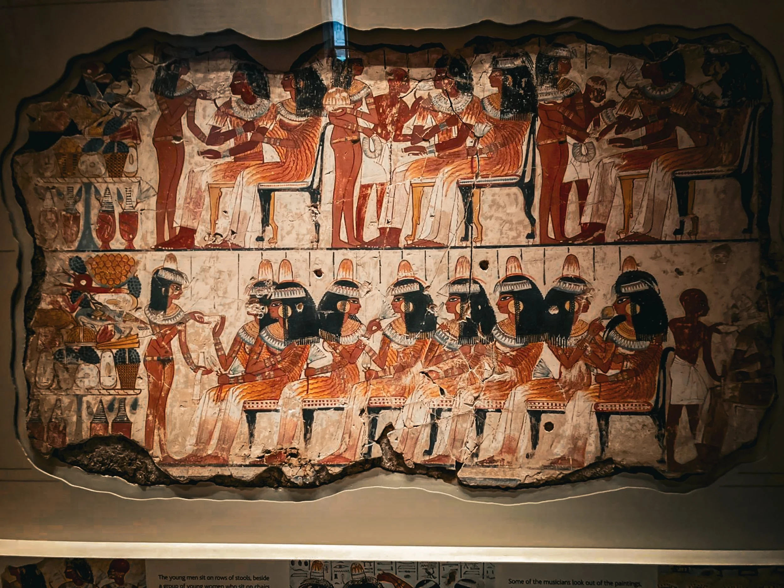 An ancient Egyptian tomb painting displayed in a museum, showing two registers of figures feasting and making music in warm ochre and black