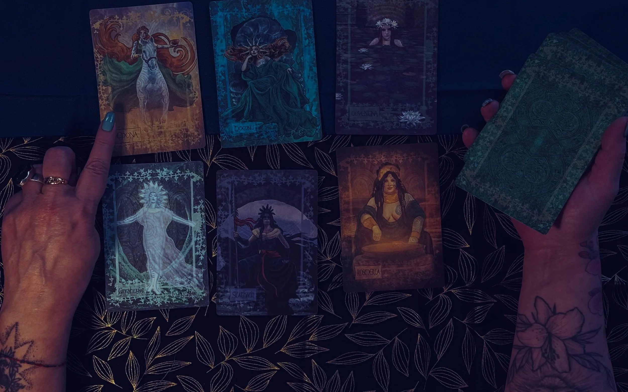 Hand of a person with tattoos holding a deck of tarot cards, with other tarot cards laid out on a dark, patterned surface.