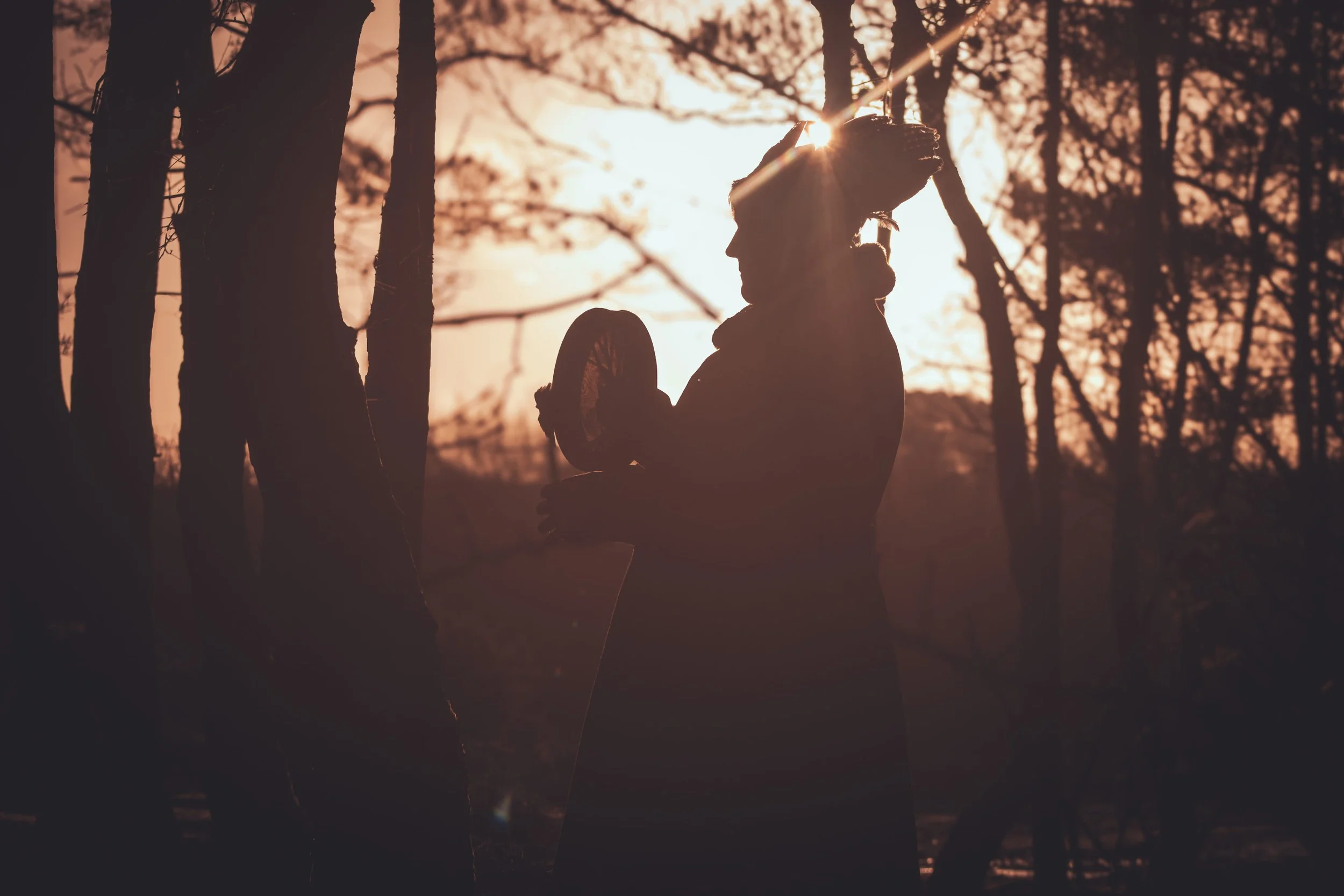 Silhouette of a woman holding a bicycle in a wooded area at sunset or sunrise.