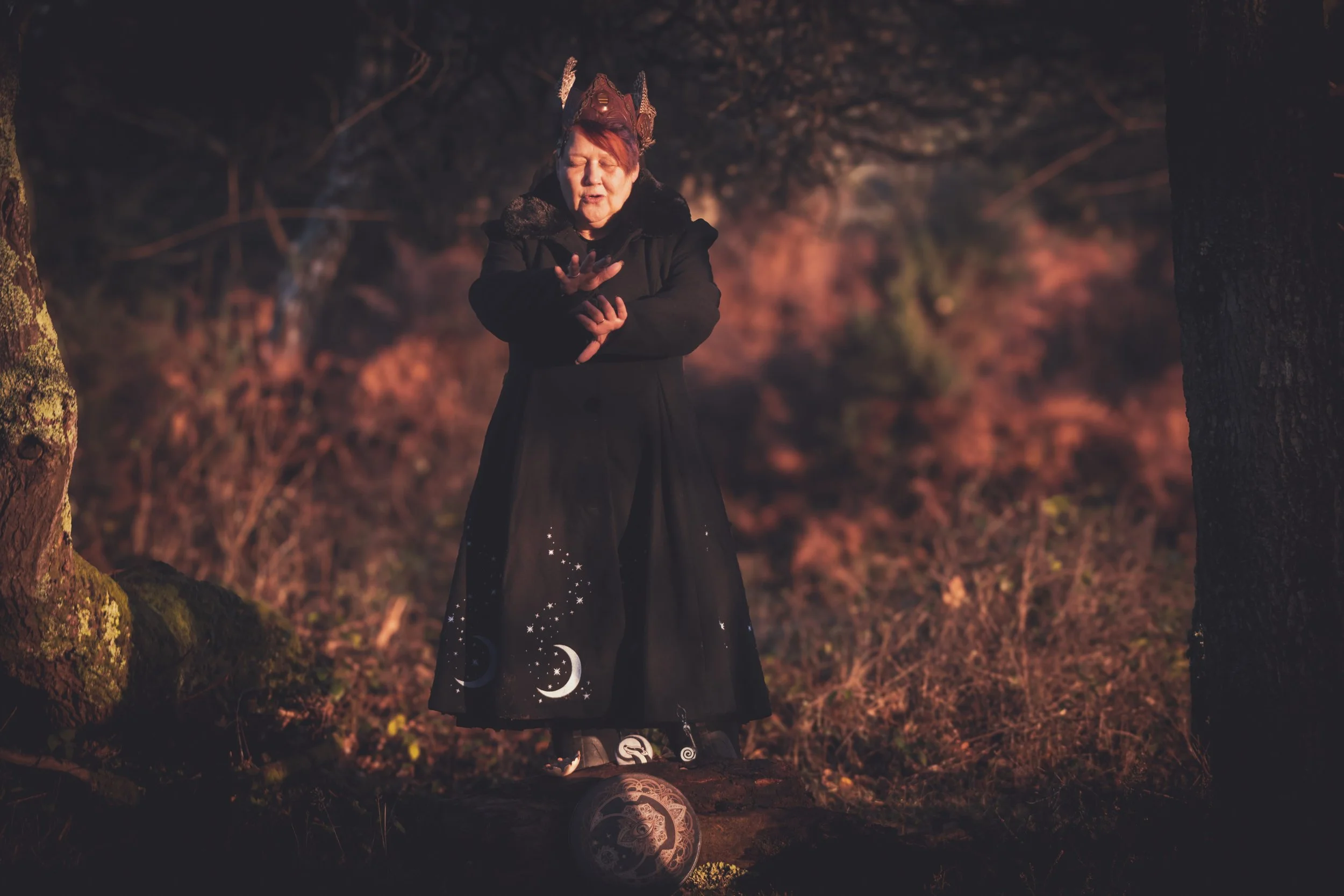 A woman dressed as a witch with a black cloak decorated with celestial symbols, and a witch hat, standing outdoors in a wooded area during twilight, with trees and fall foliage in the background.